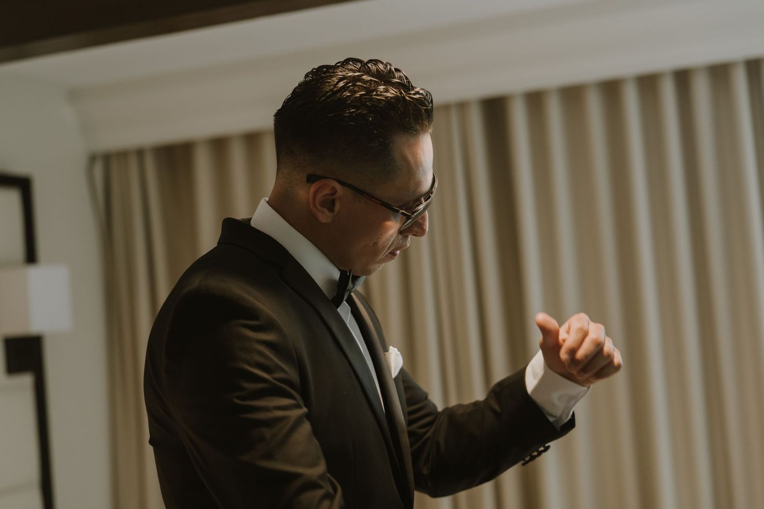 Groom in formal black tuxedo and sunglasses adjusting cuff in indoor setting.