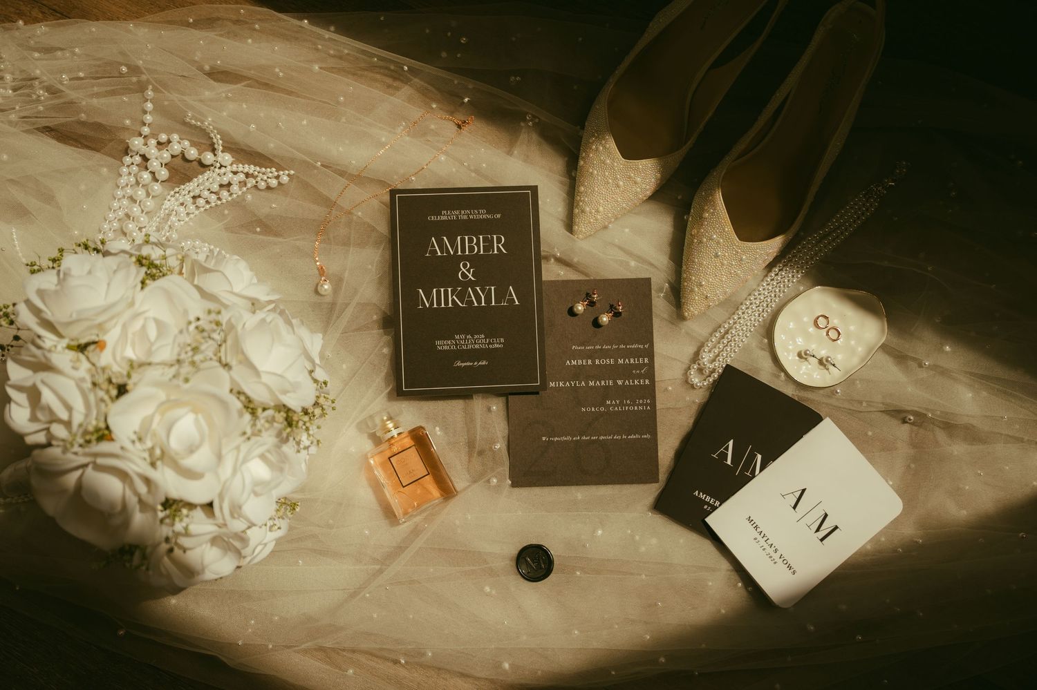 Elegant wedding flat lay with floral bouquet, dark invitations for Amber & Mikayla, perfume, earrings, and heels.