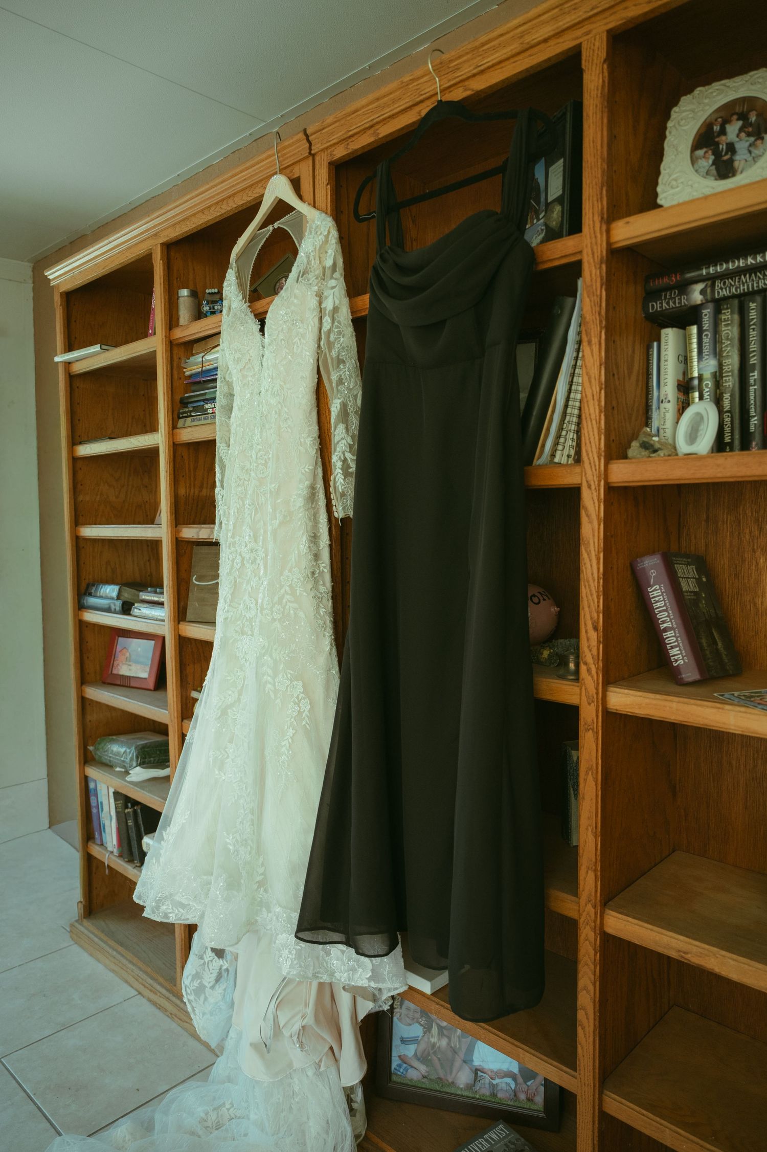 Wedding dress and dark bridesmaid gown hanging on a wooden bookshelf before the ceremony.