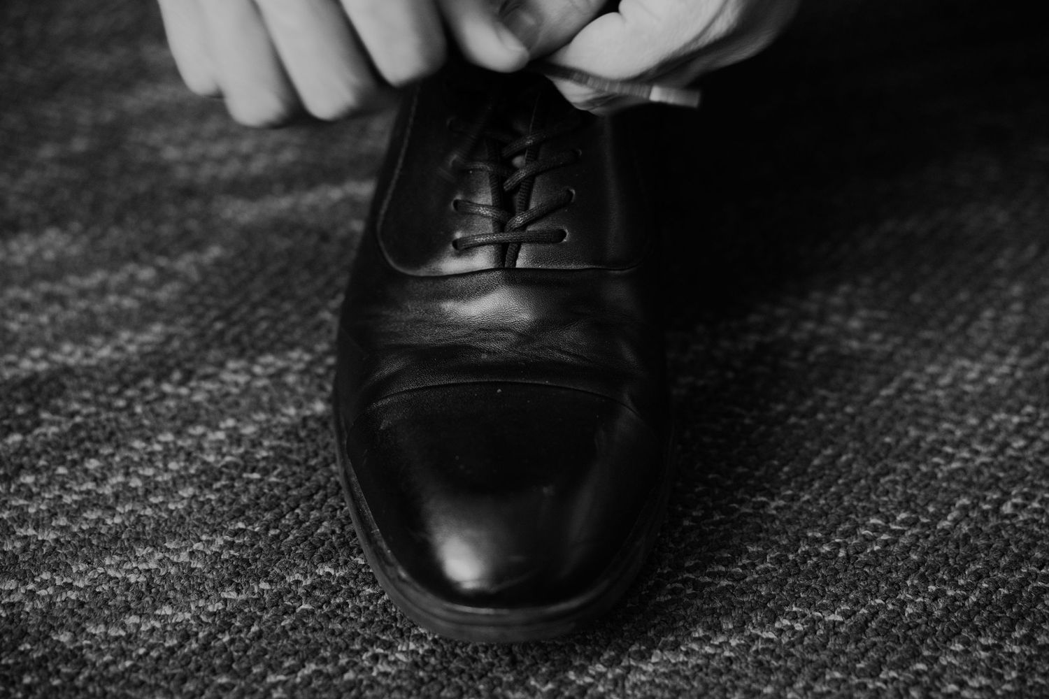Black and white photo of hands tying laces on a polished black Oxford dress shoe on carpet.