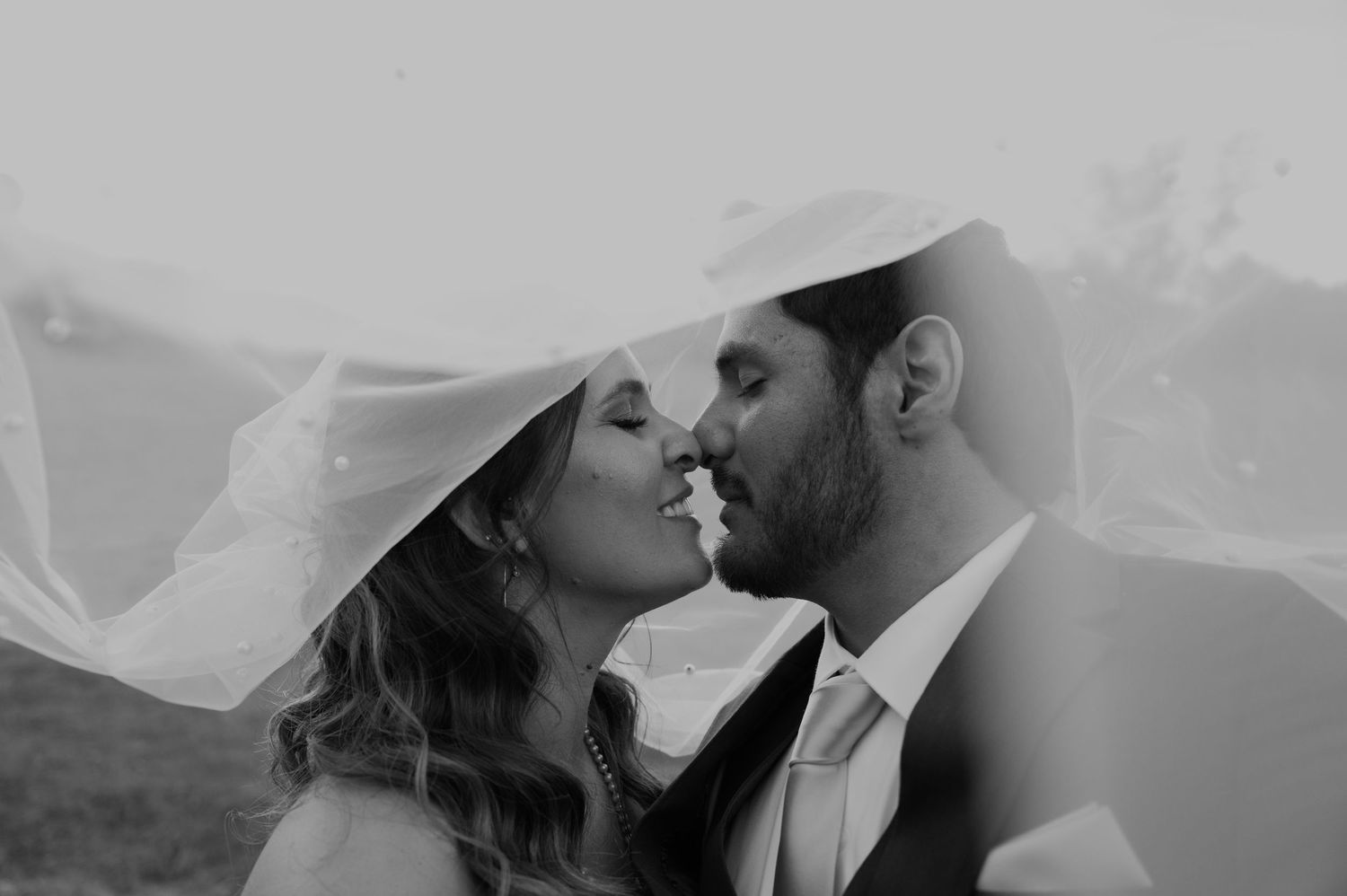 Black and white photo of a bride and groom sharing a romantic moment under a flowing wedding veil.