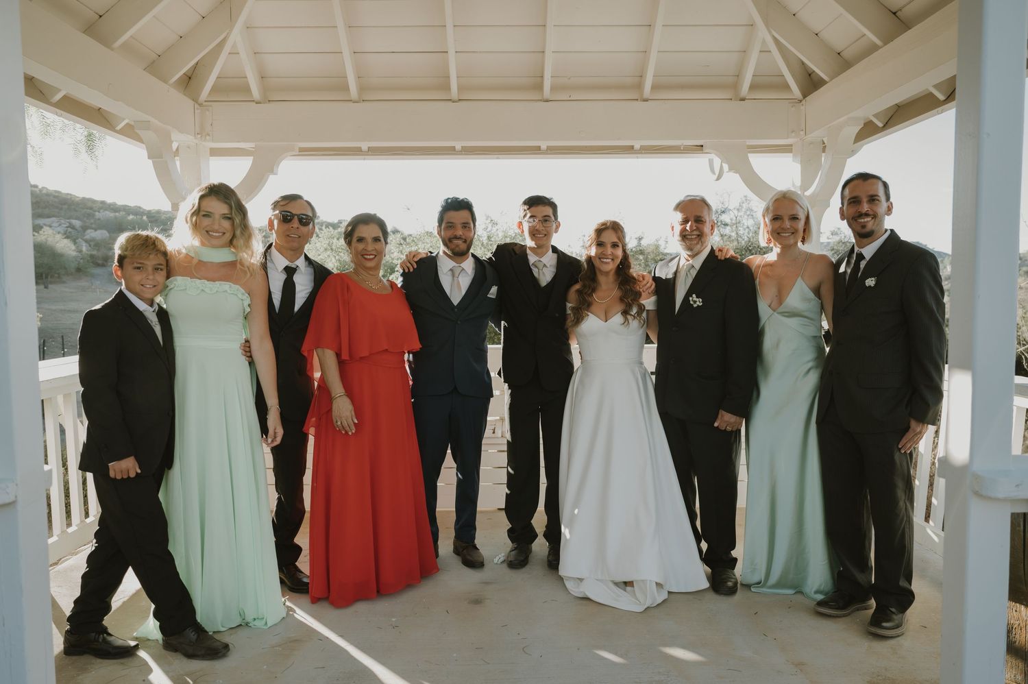 Wedding party poses together under white gazebo in mix of formal attire and bridesmaid dresses.