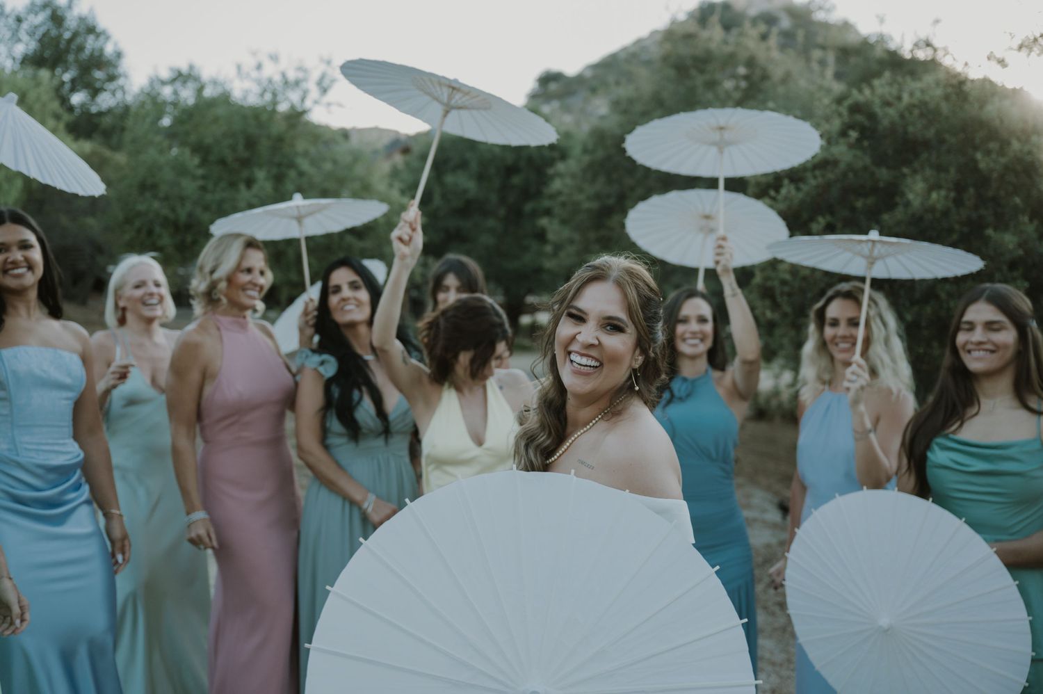 Smiling bride poses with bridesmaids in colorful dresses holding white parasols outdoors among green trees.