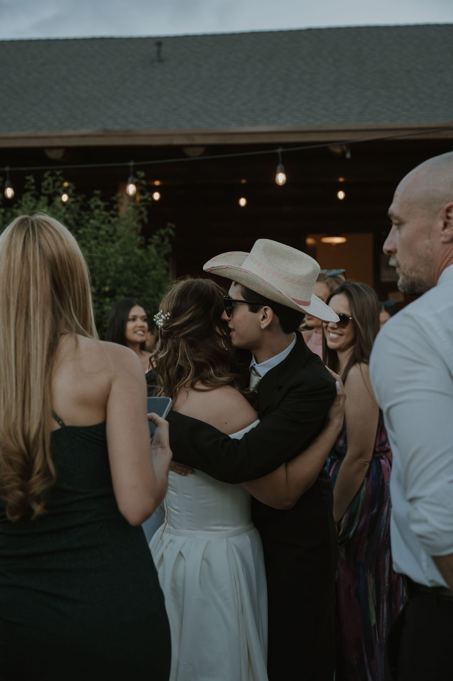 Bride and groom share a kiss while dancing at an outdoor wedding reception with string lights.