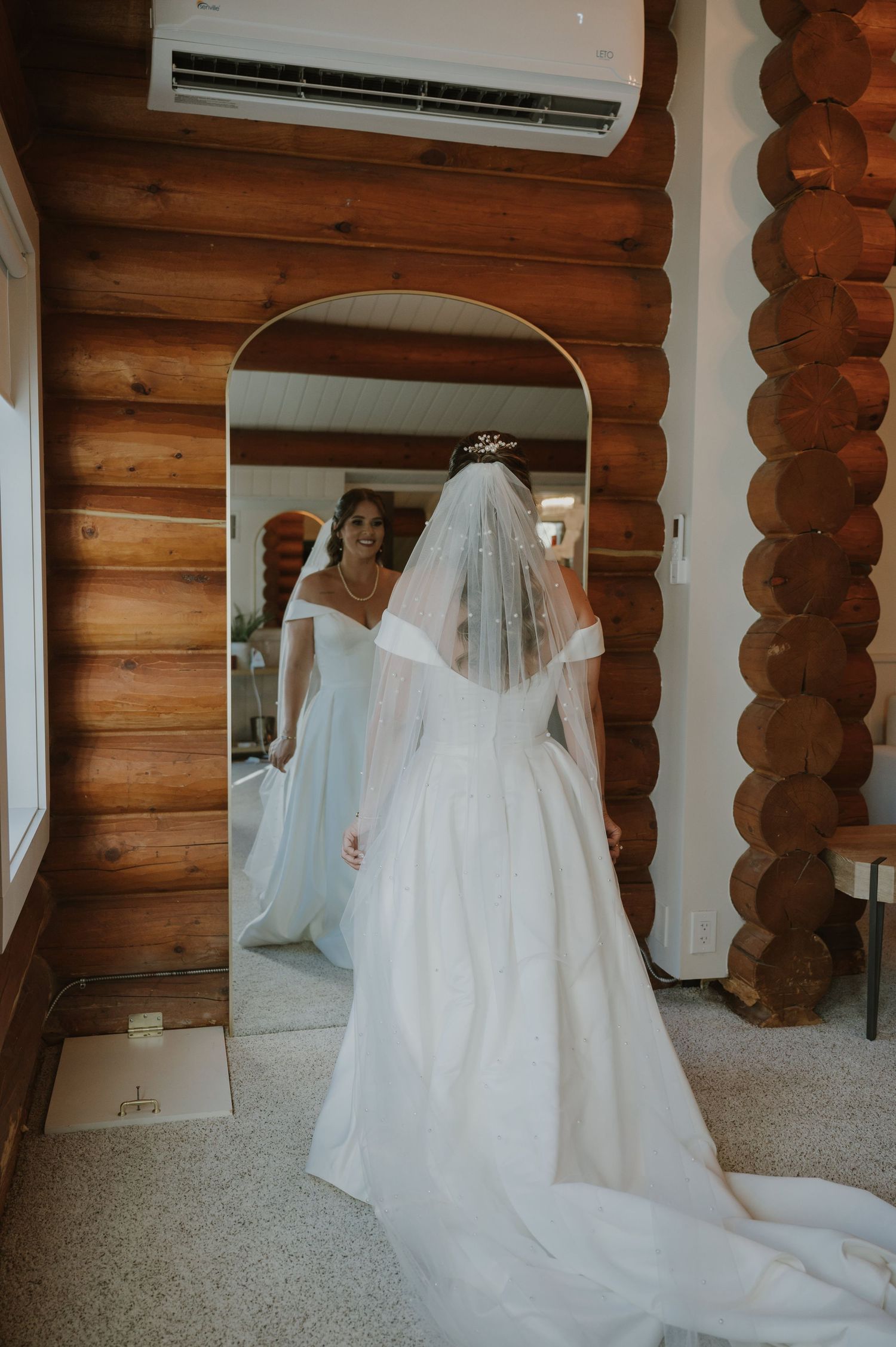 Bride in white off-shoulder gown and veil stands before arched mirror in rustic log cabin bridal suite.