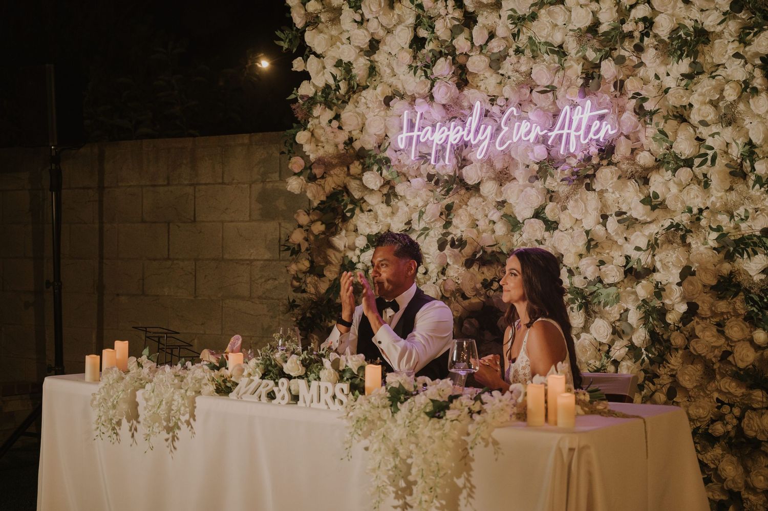 Couple seated at floral sweetheart table under Happily Ever After neon sign at elegant outdoor wedding reception.