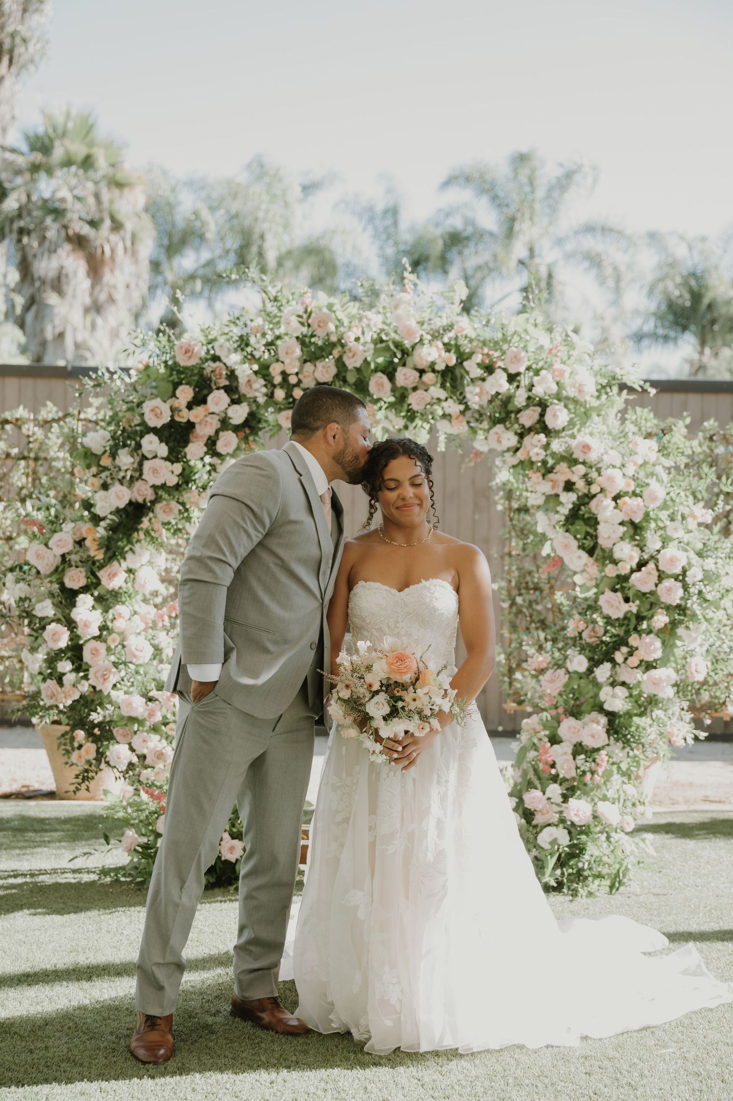 Bride and groom share a romantic moment under a floral arch adorned with pink roses at an outdoor wedding ceremony.
