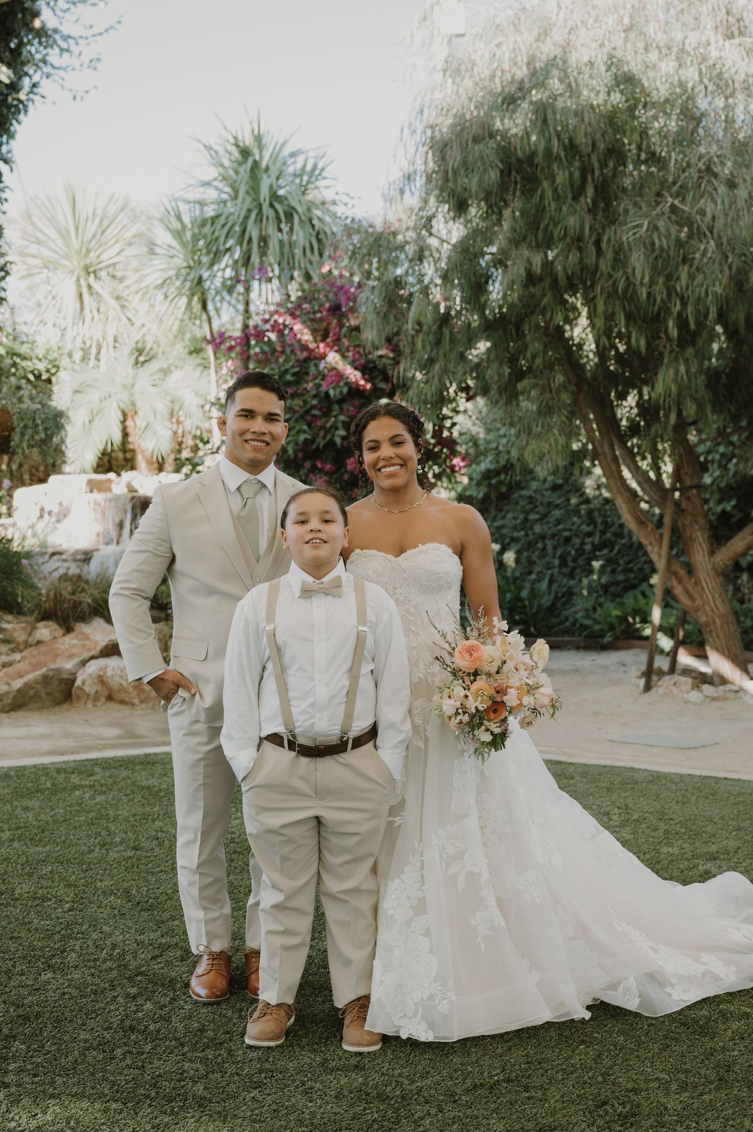 Bride and groom pose with young ring bearer in garden at outdoor wedding ceremony.