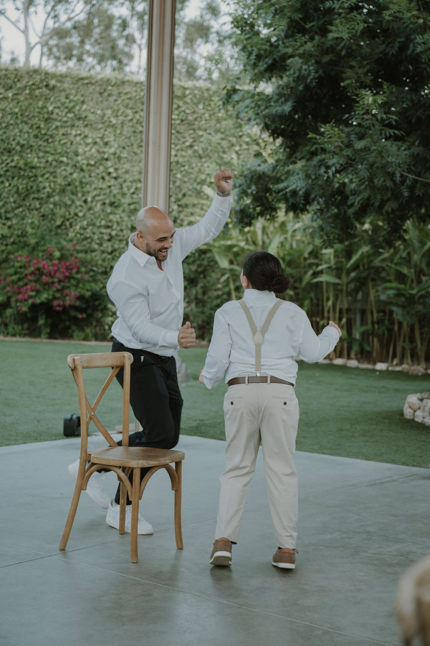 A man and boy in white shirts dance joyfully outdoors at a wedding reception on a patio surrounded by greenery.