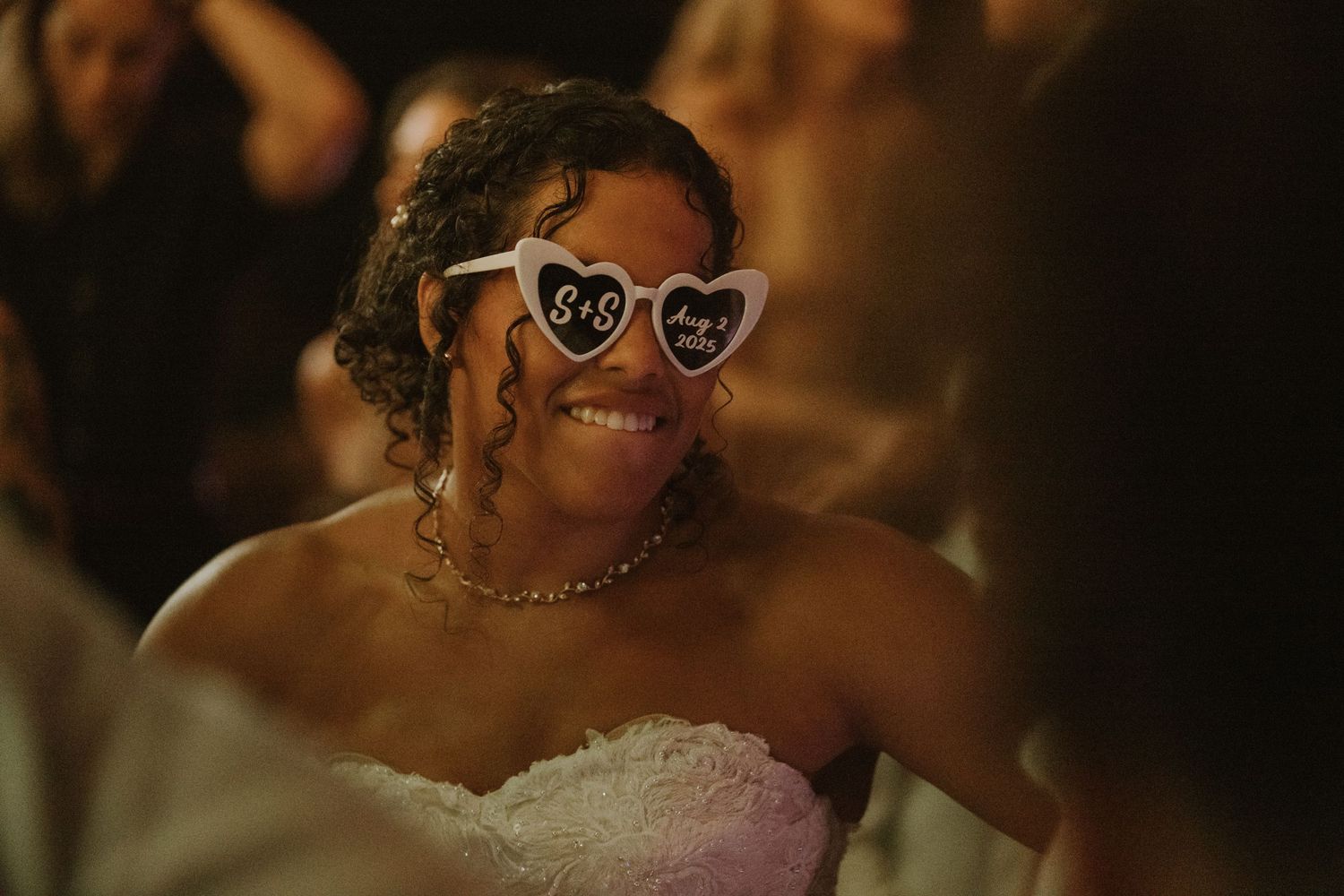 Smiling bride wearing heart-shaped novelty sunglasses with initials at a wedding reception in a strapless white gown.