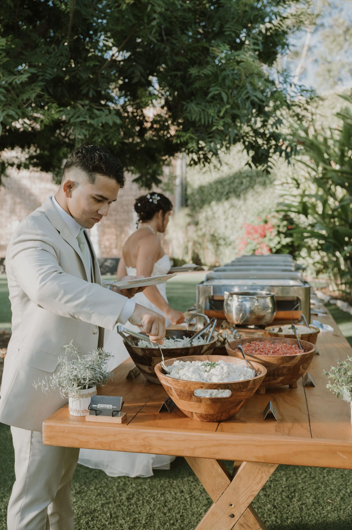 Groom in beige suit serving food at outdoor wedding buffet table with bride in background.