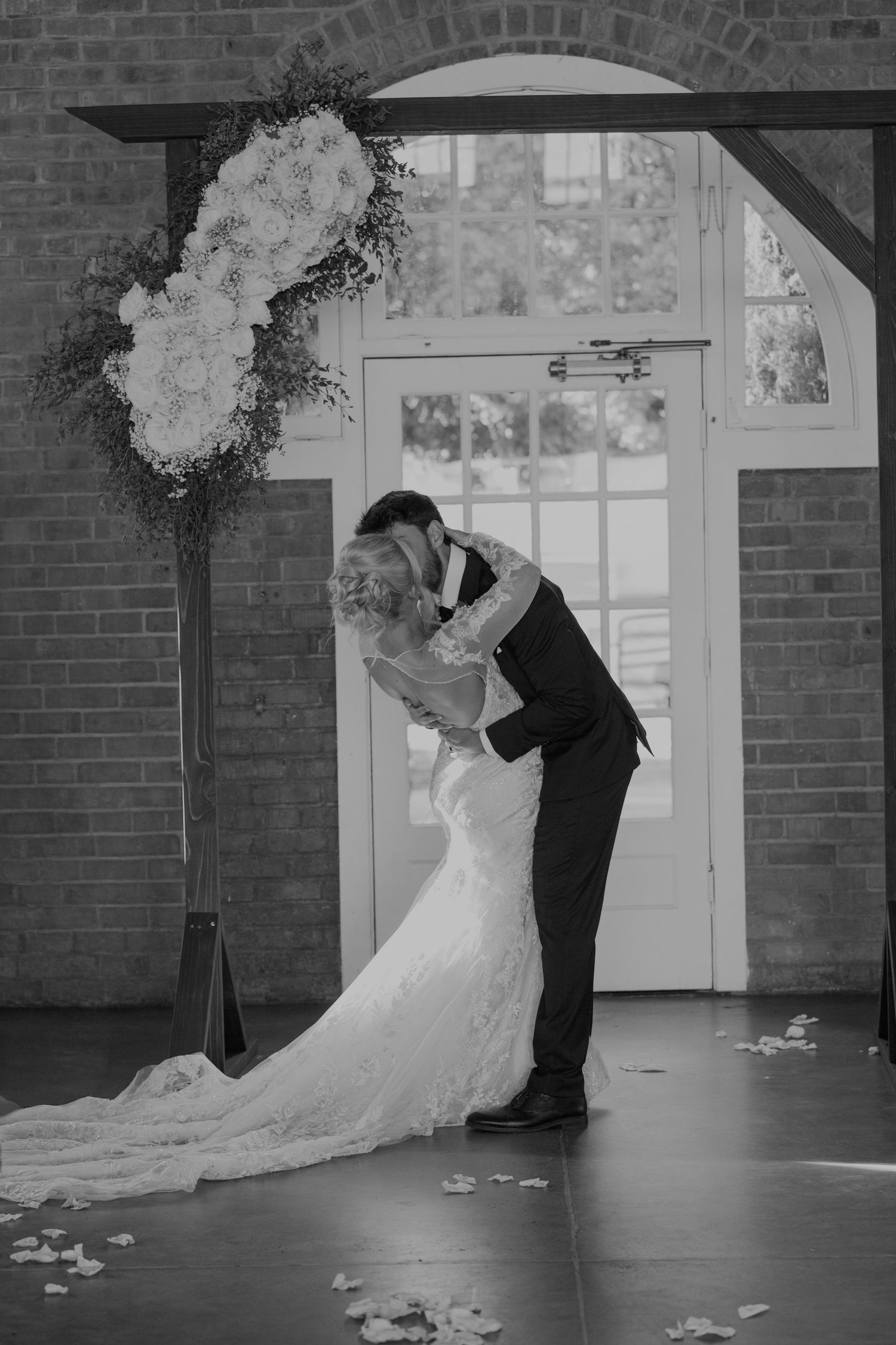 Black and white photo of bride and groom kissing under floral arch at wedding ceremony.