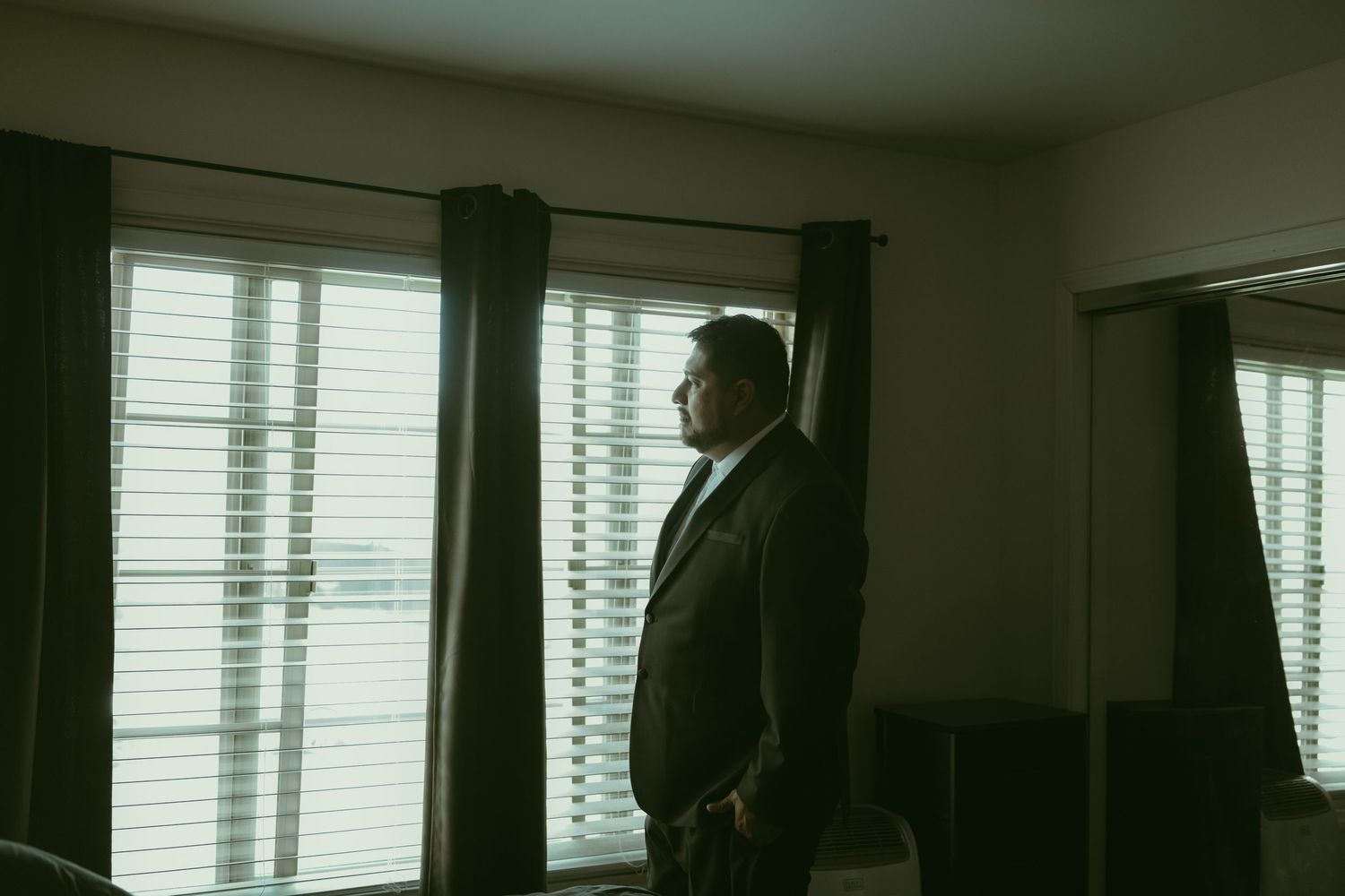 Groom in dark suit standing by window with blinds in dimly lit room.