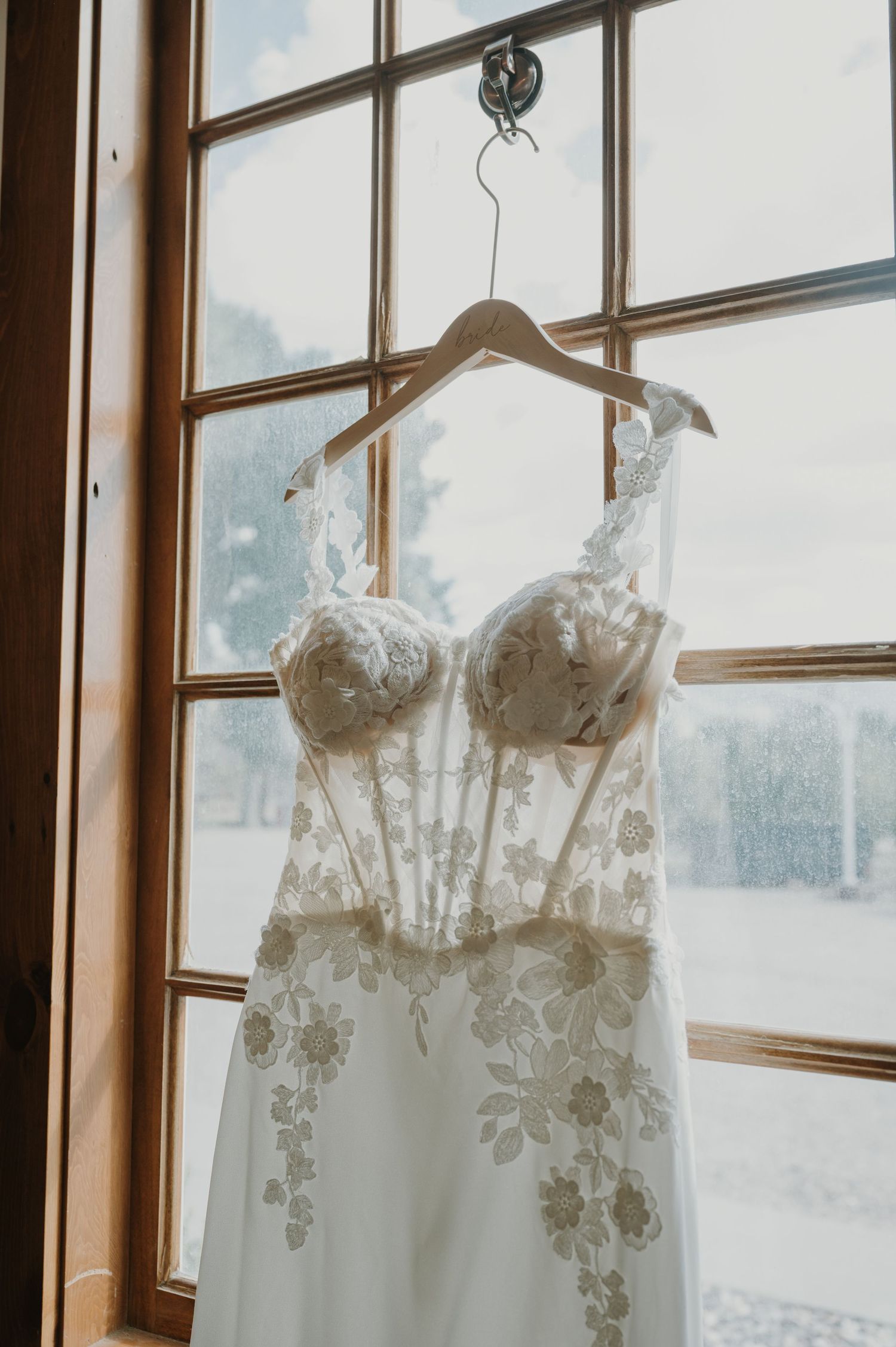 White lace wedding dress with floral details hangs by a window in natural light.