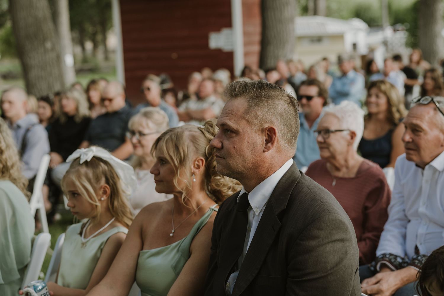 Wedding guests seated in rows watch the ceremony proceedings under tall trees.