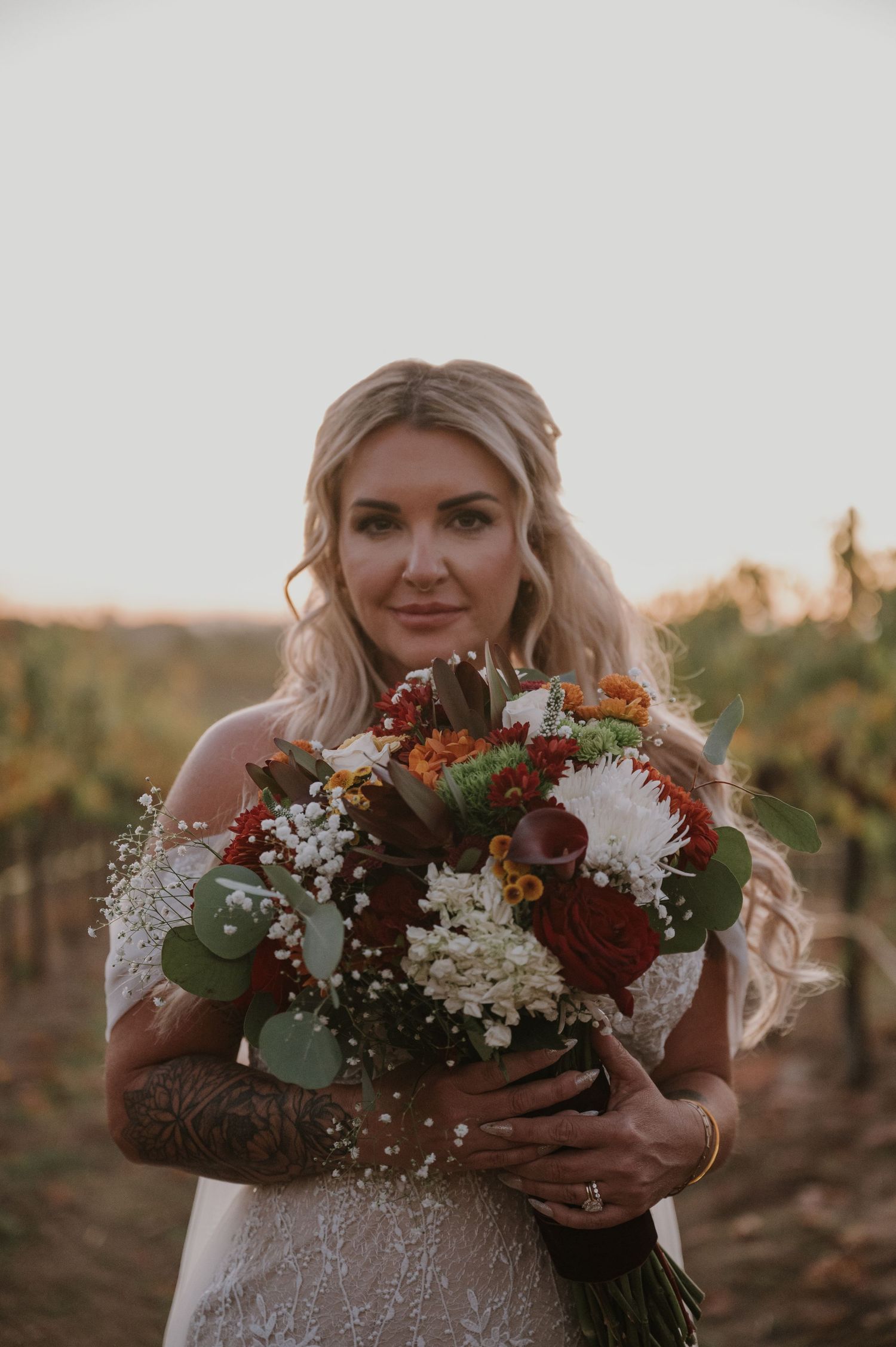 A bride holds a vibrant autumn bouquet with burgundy and white flowers in a vineyard setting.