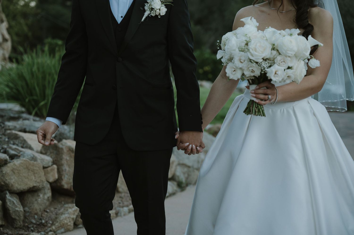 Newlywed couple holding hands with bride in white ballgown and groom in black suit holding white bouquet.