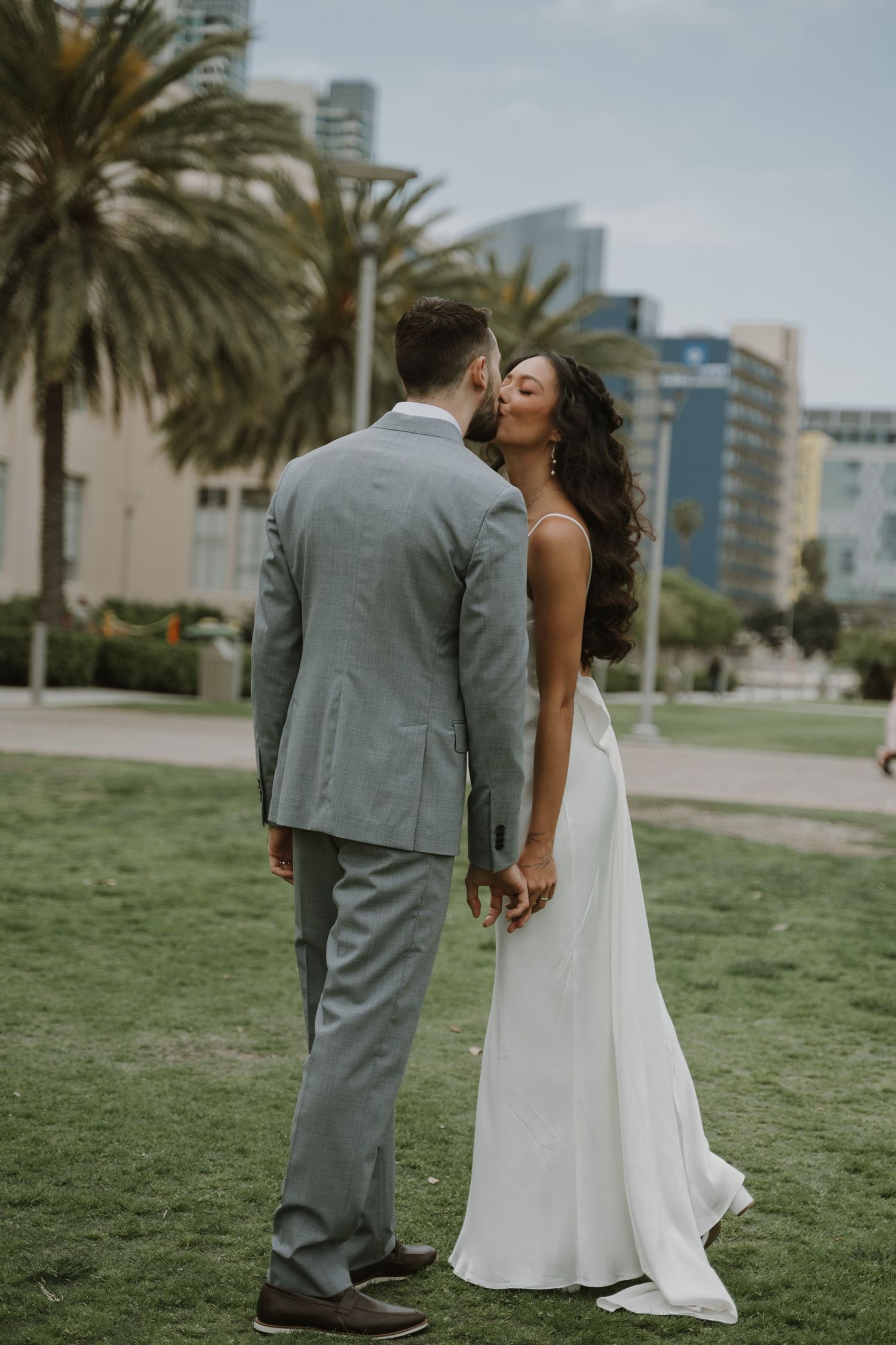 Wedding couple shares playful moment on grassy lawn with palm trees and city skyline.