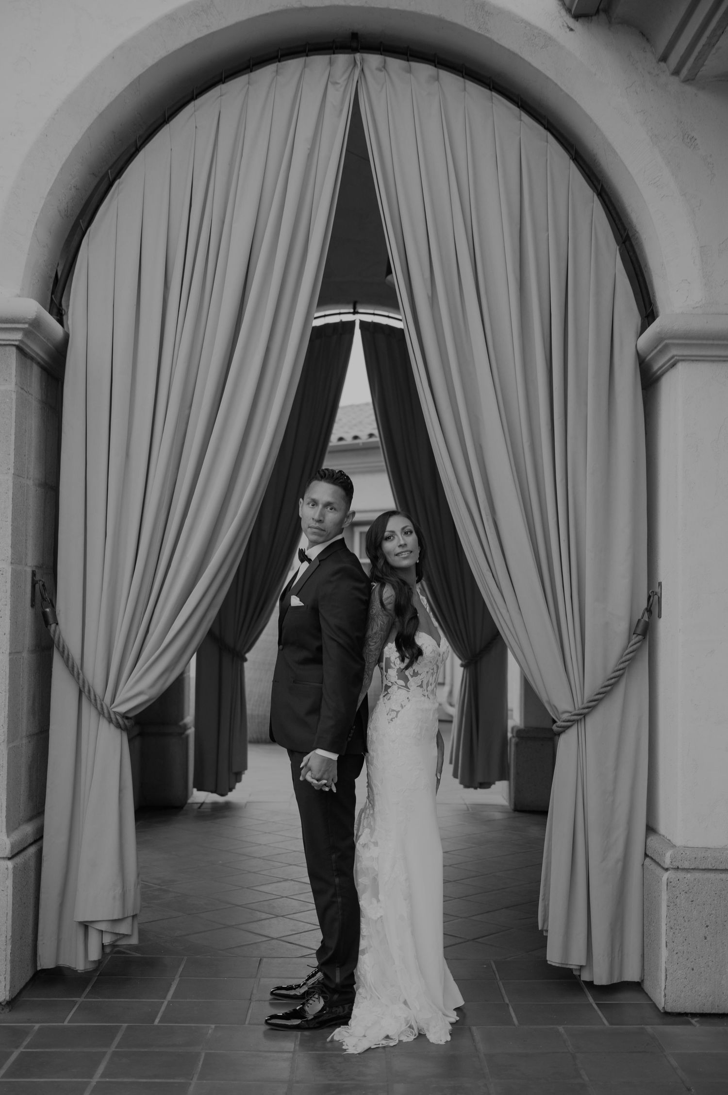 A black and white photo of newlyweds in an elegant colonnade with draped curtains.