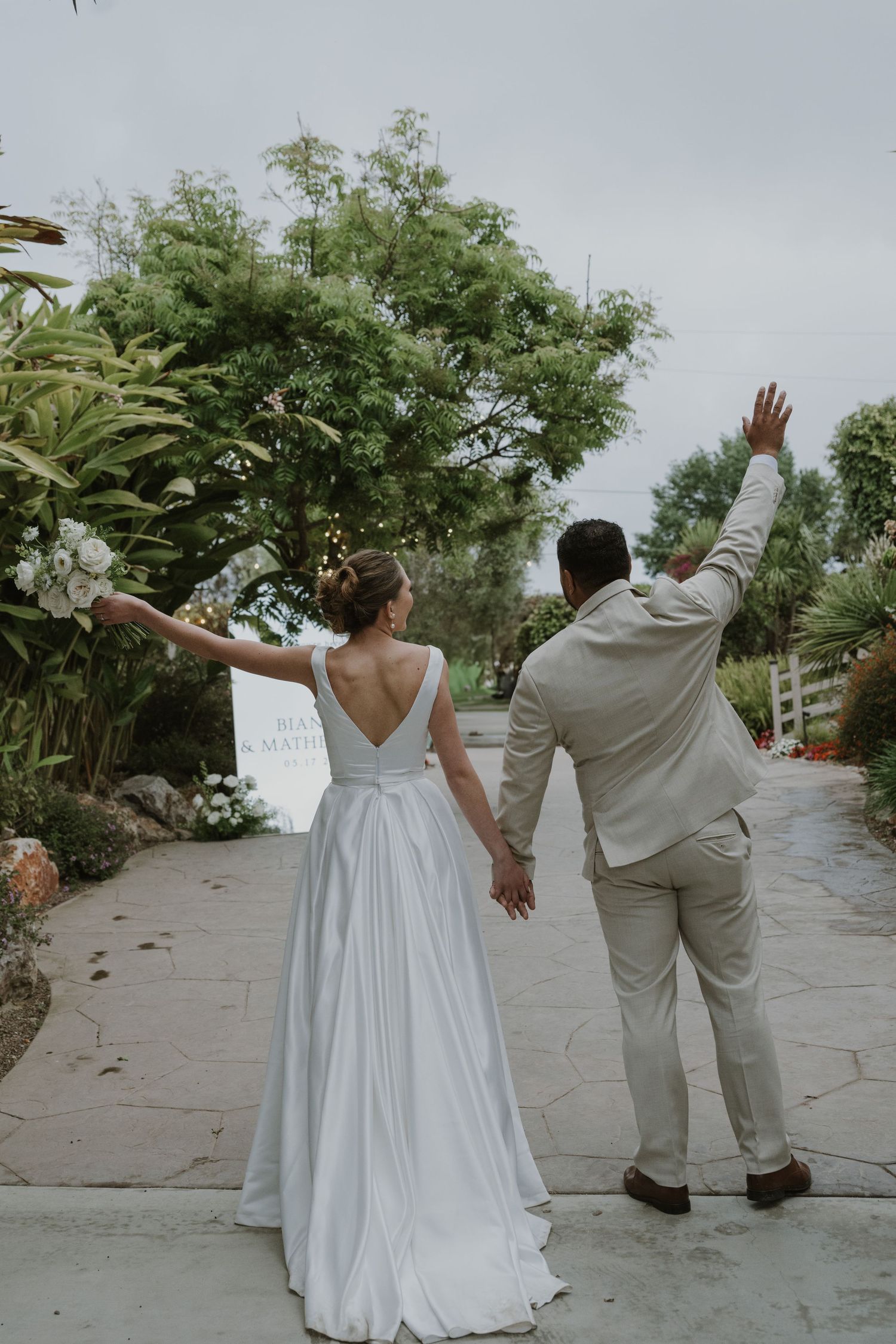Bride in white gown and groom in tan suit hold hands, waving as they walk through a lush garden path.