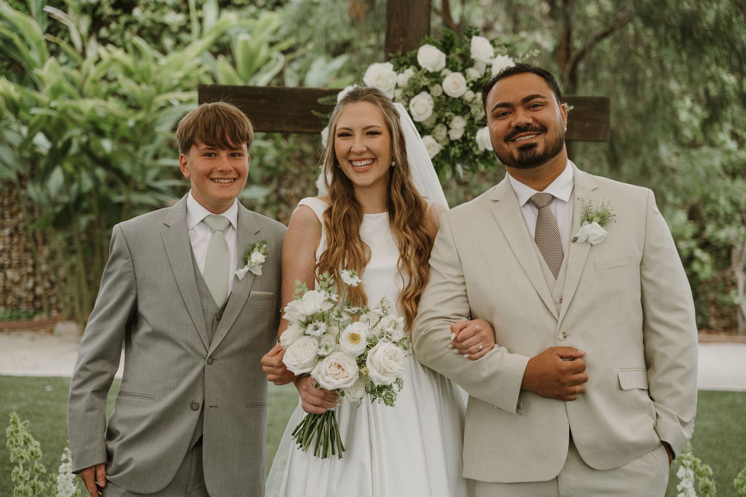 Bride in white gown holds bouquet flanked by groom in beige suit and groomsman in gray at outdoor wedding.