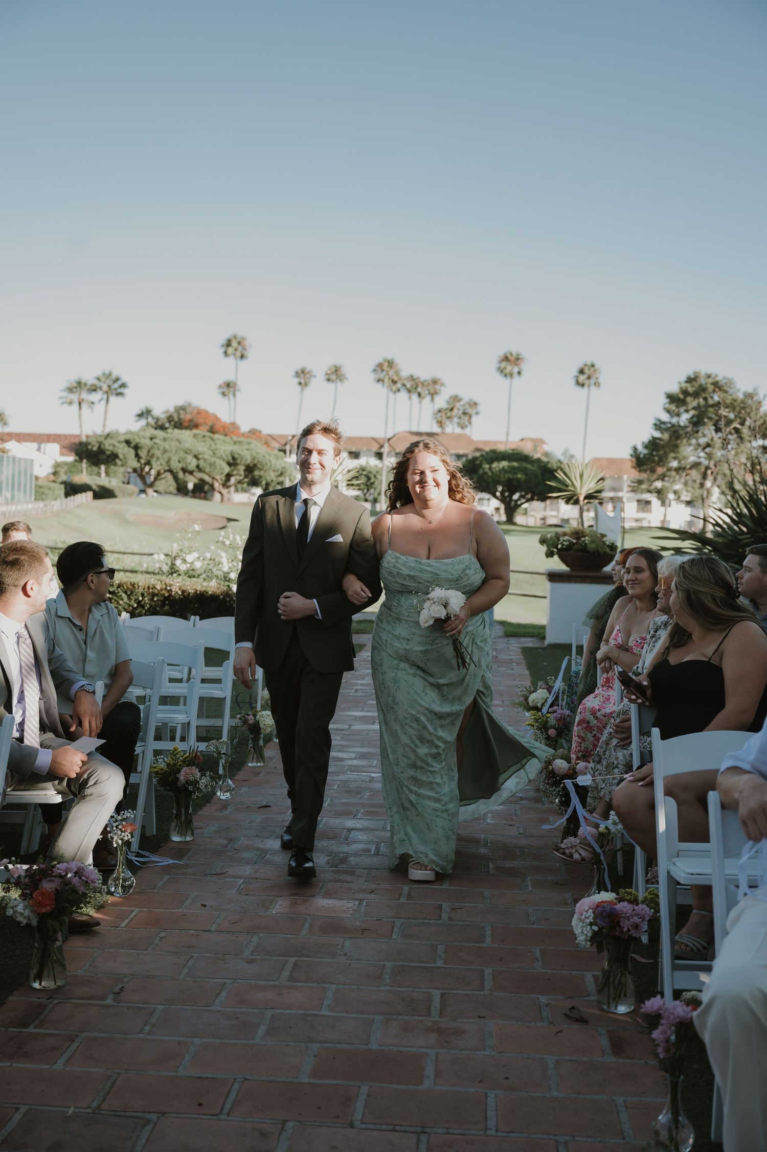 Bride and groomsman walk down brick aisle at outdoor wedding ceremony with palm trees and white chairs.