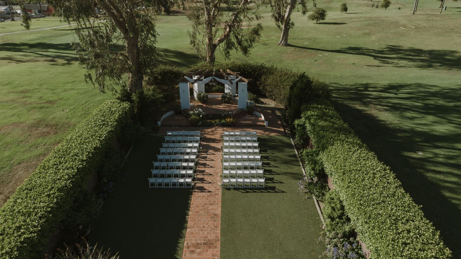 Aerial view of an elegant outdoor wedding ceremony setup with white chairs, hedges, and a decorative arch on lush green grounds.
