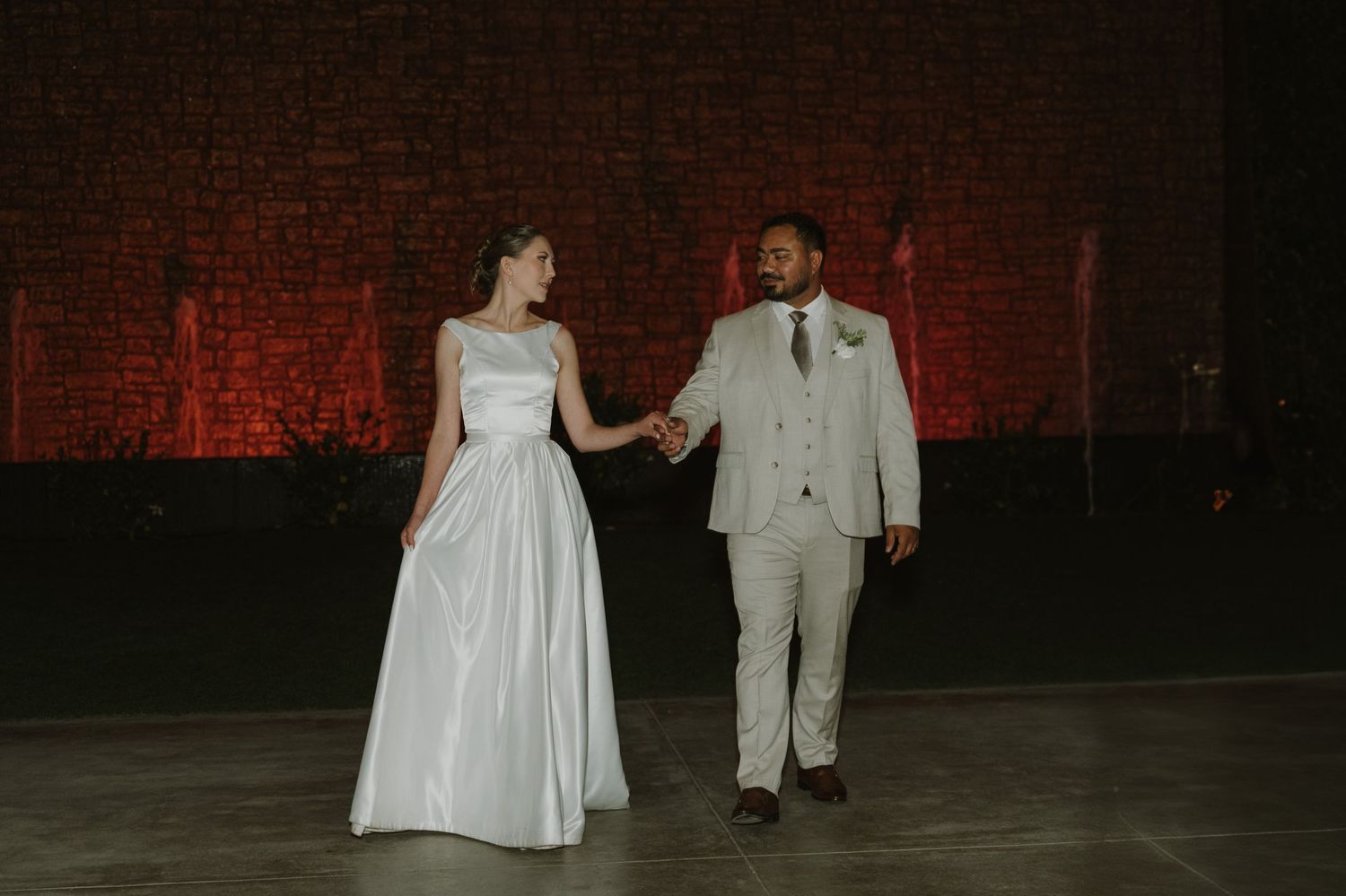 Bride and groom hold hands smiling during their first dance at a romantic wedding reception with red brick backdrop.