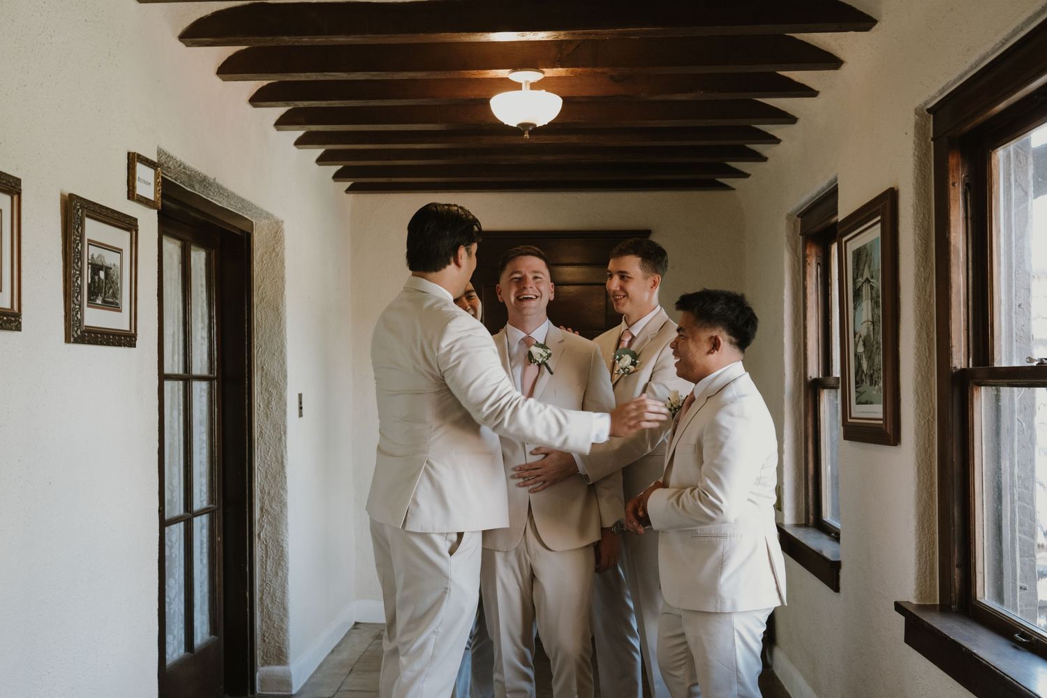 Groomsmen in beige suits chatting in a sunlit hallway with wooden beam ceiling before a wedding ceremony.