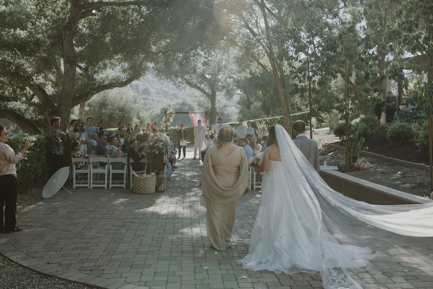 Bride in white gown and veil walks toward outdoor wedding ceremony under lush trees on a brick pathway.