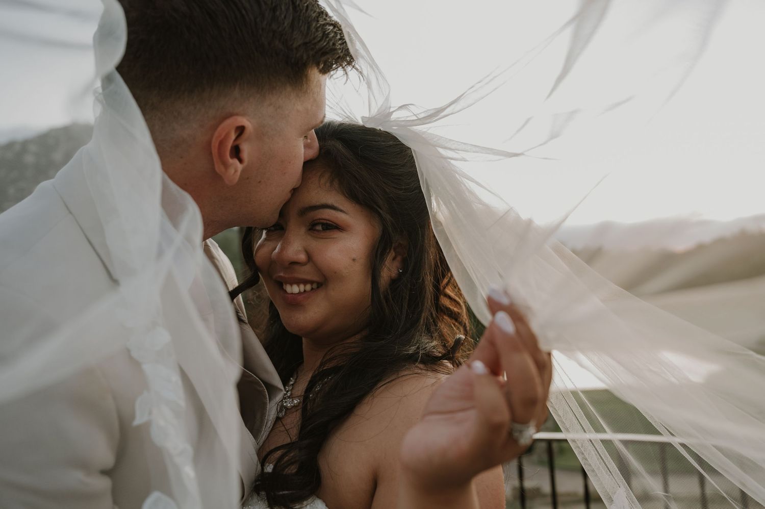 Bride smiles joyfully as grooms veil wraps around them in a candid wedding portrait.