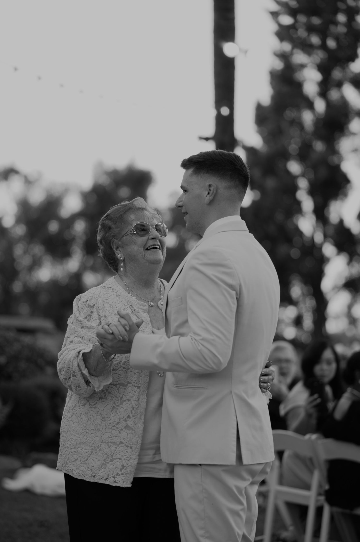 Groom in white suit sharing a tender mother-son dance at outdoor wedding reception in black and white photo.