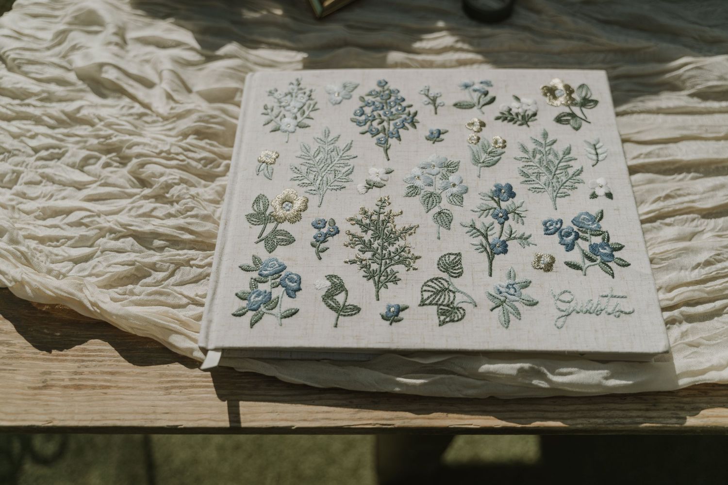 A botanical illustrated journal with blue floral designs rests on a wooden surface with white linen.