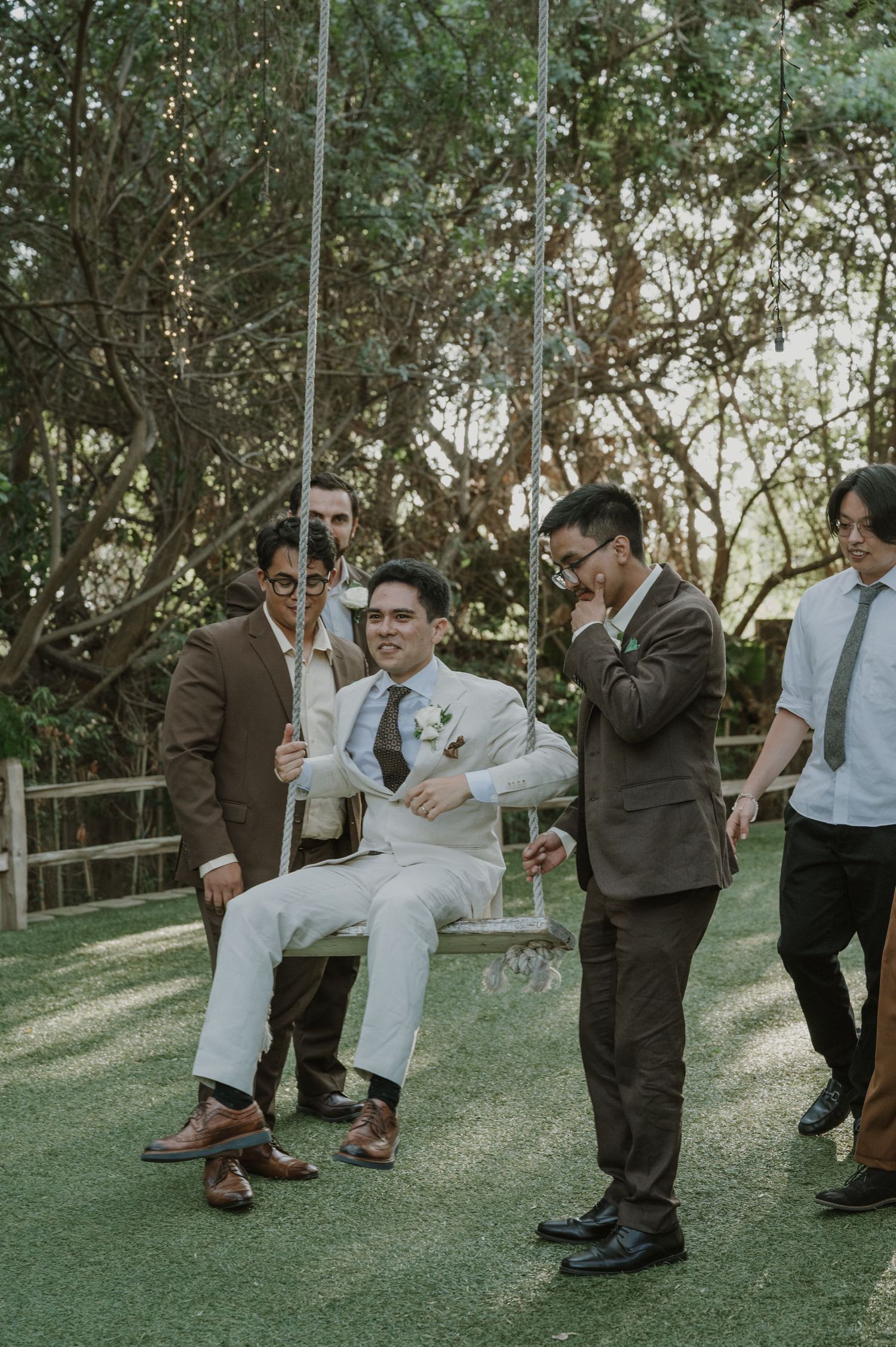 Groom in white suit sits on garden swing surrounded by smiling groomsmen in brown suits outdoors.