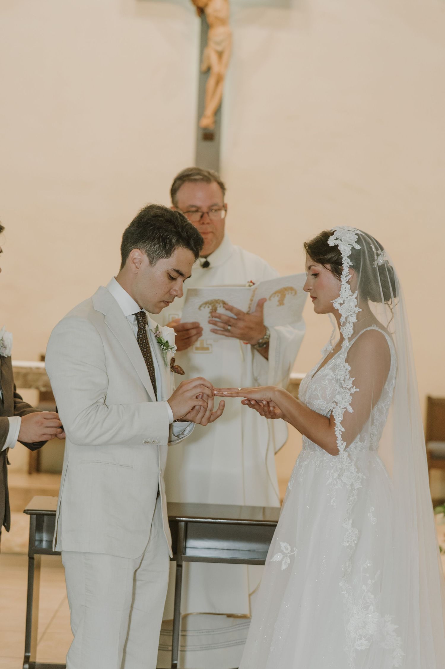 Groom in white suit exchanges rings with bride in lace veil at church altar with priest officiating.