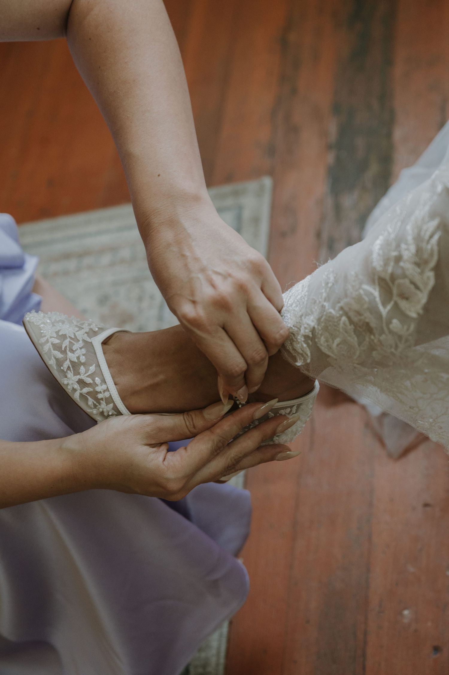 Bridesmaid helping bride put on white lace wedding shoe, close-up detail shot on wooden floor.