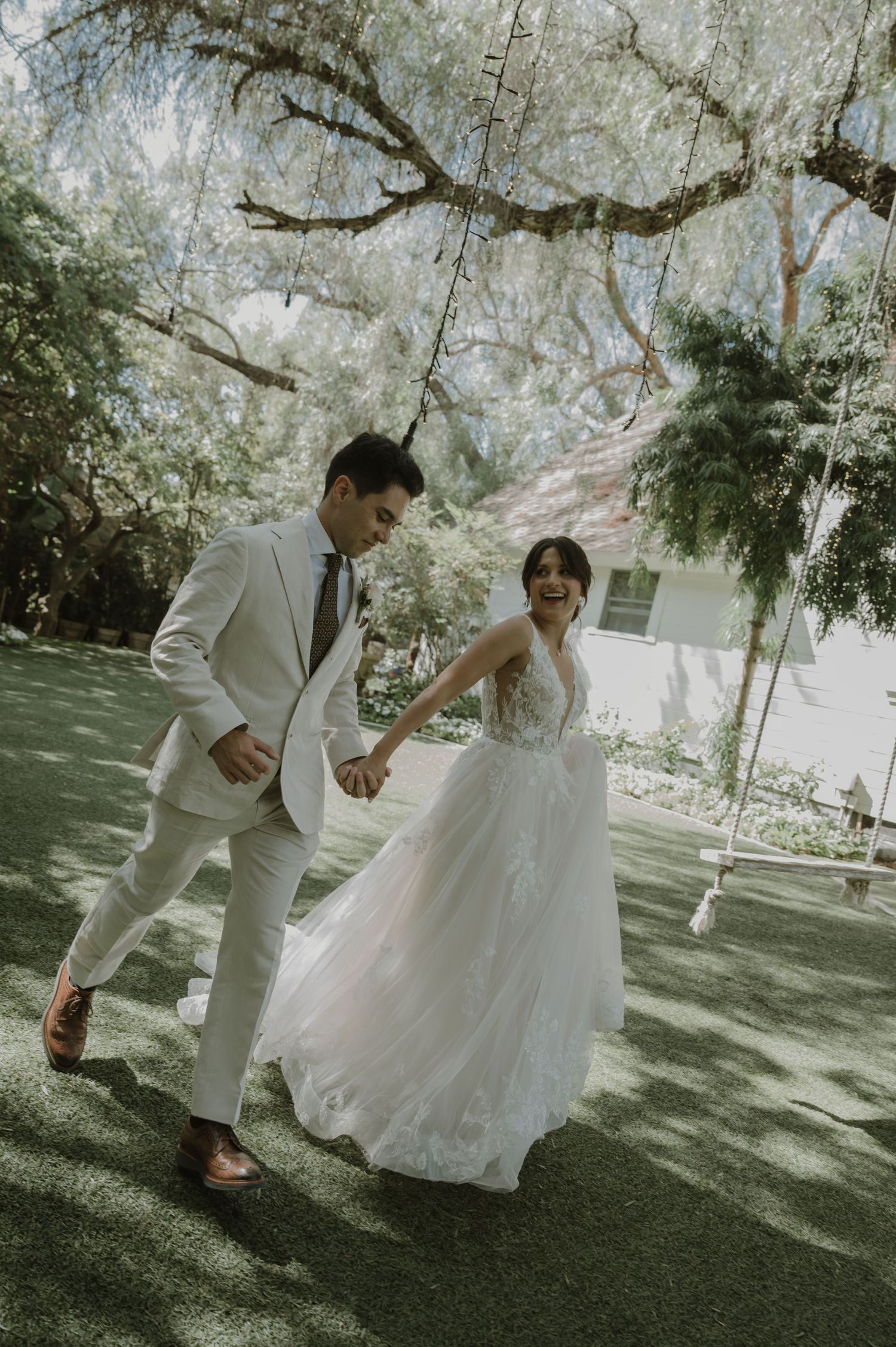 A joyful bride in a flowing white lace gown and groom in a cream suit hold hands and dance outdoors under lush trees.