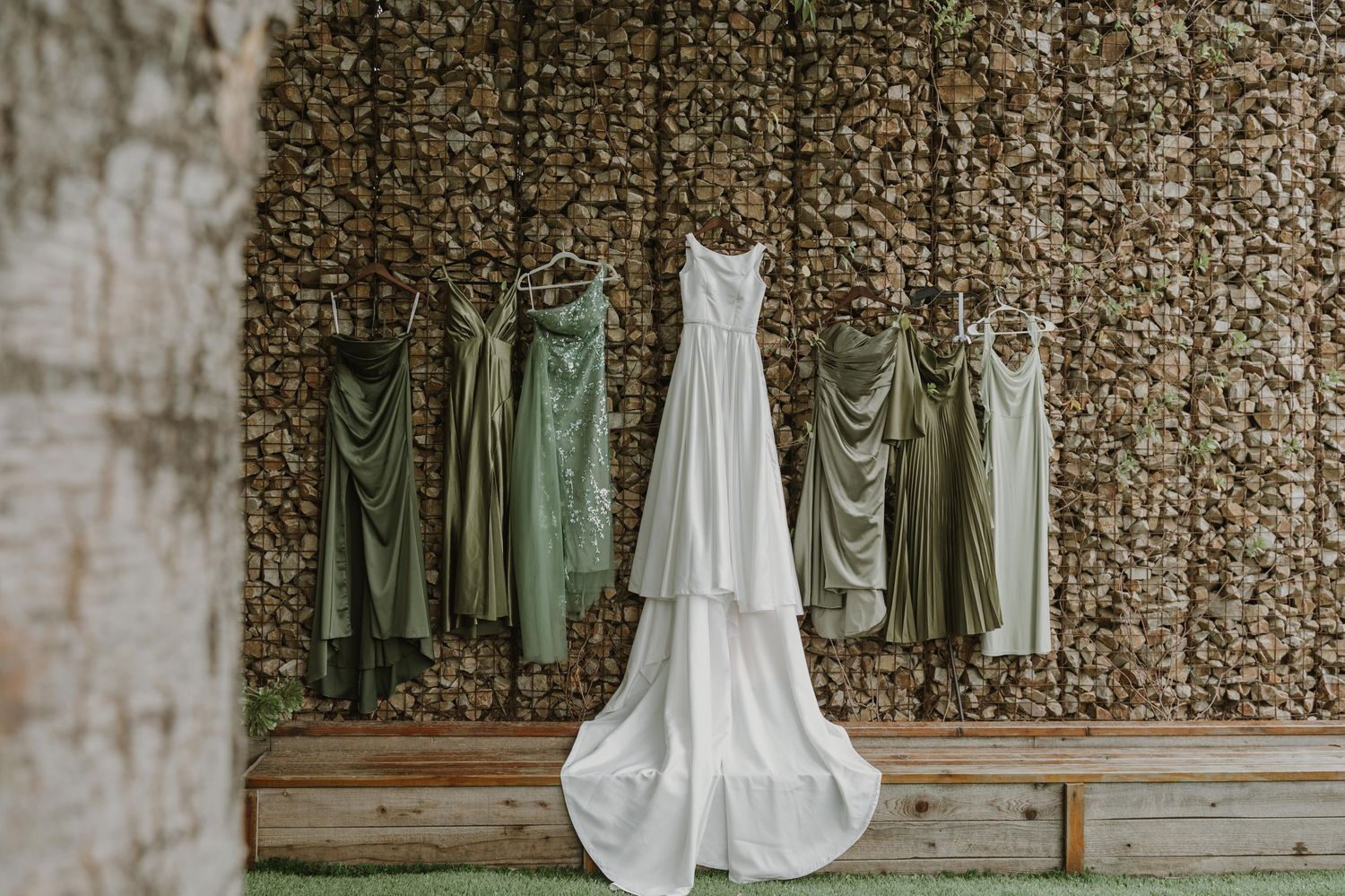 White wedding gown and sage green bridesmaids dresses hanging against a rustic stone wall backdrop outdoors.