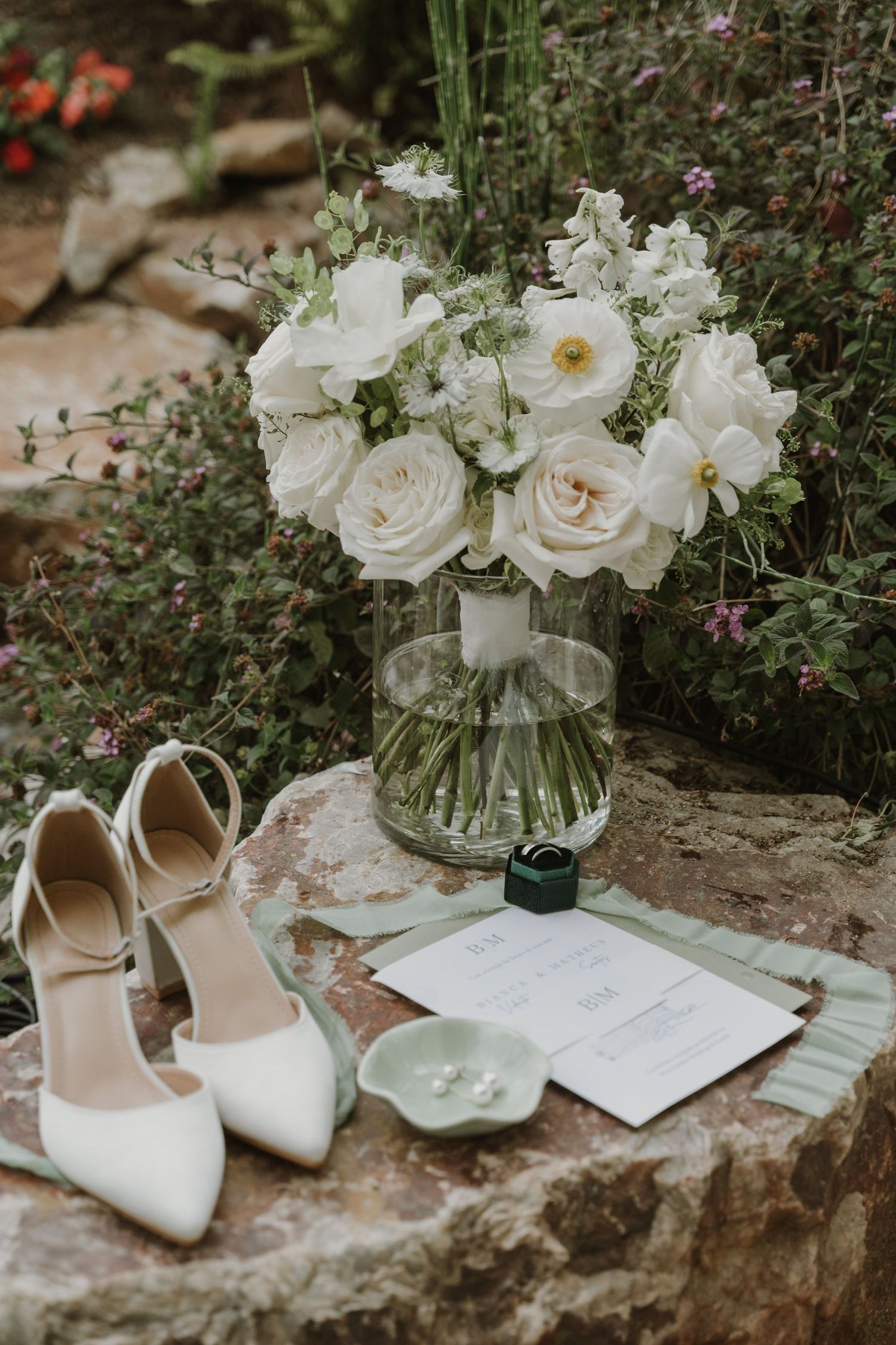 Elegant wedding flat lay with white floral bouquet, bridal heels, ring box, and stationery on stone surface.