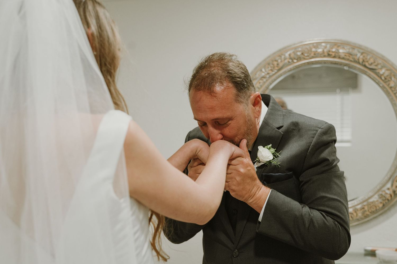 Emotional father kissing bride's hand during first look moment in front of ornate mirror.