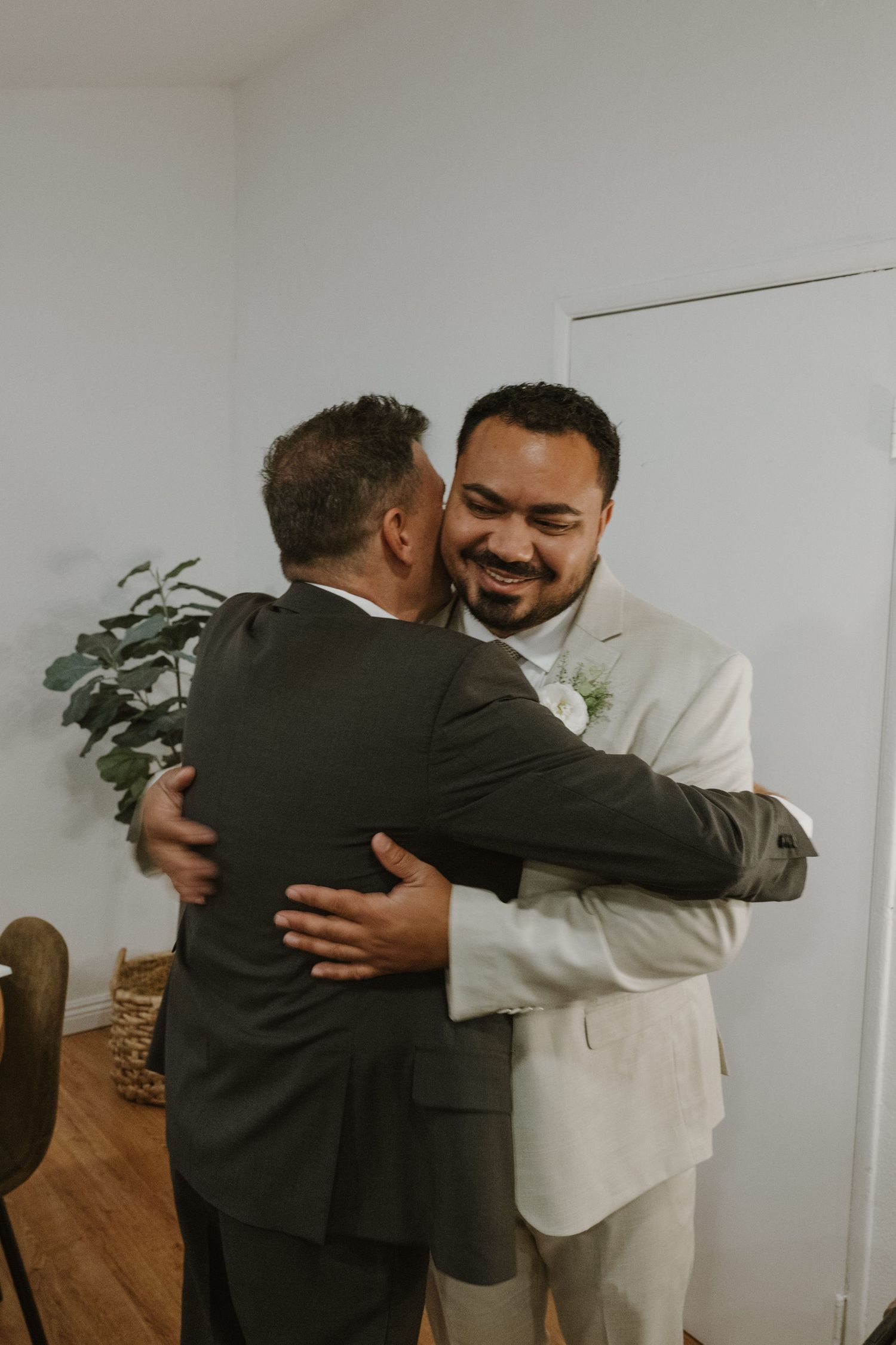 Two men in suits share a warm embrace, one wearing a light beige jacket with a floral boutonniere at a wedding celebration.