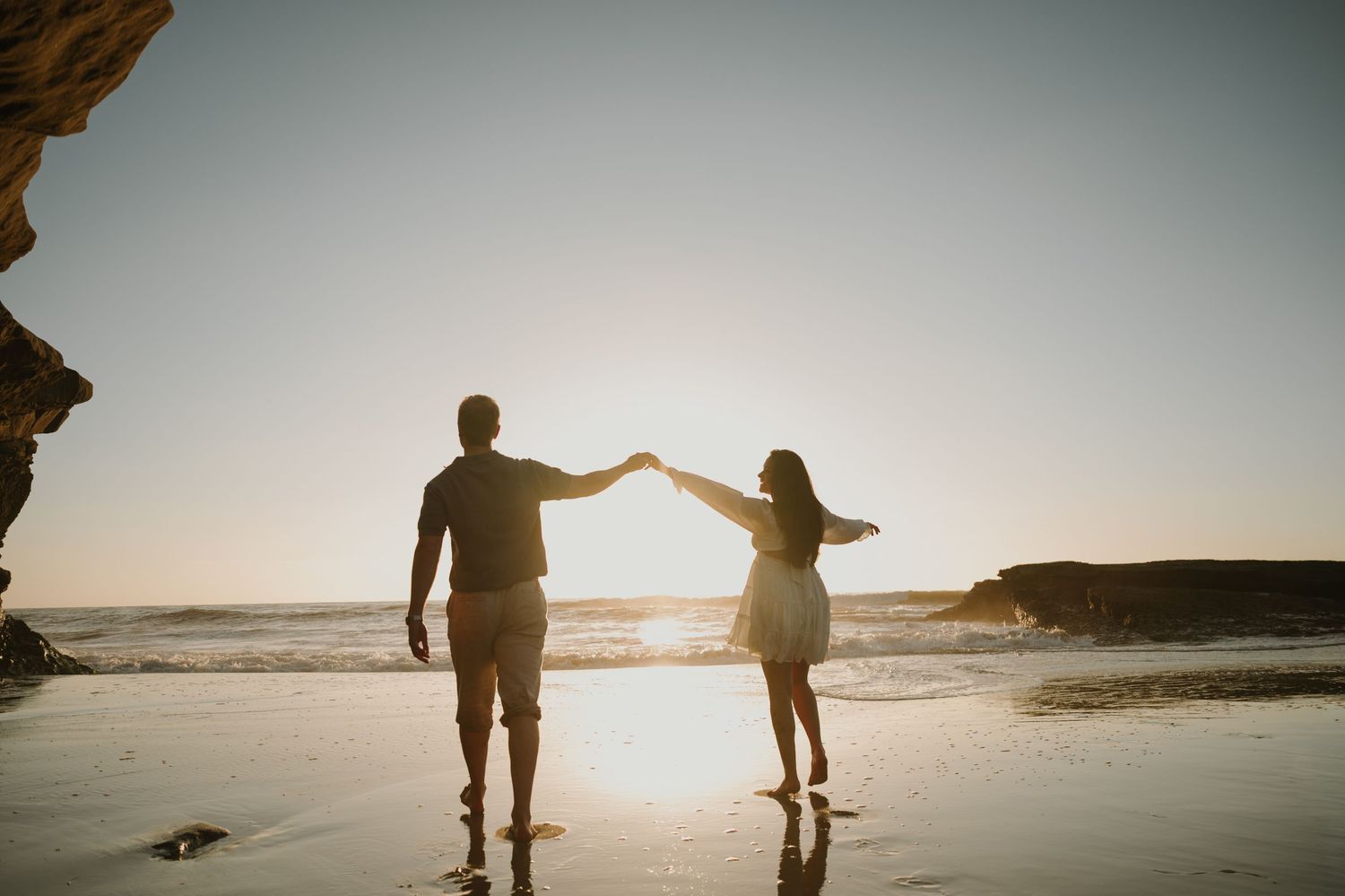 Couple holding hands dancing on a sandy beach at sunset with ocean waves in the background.