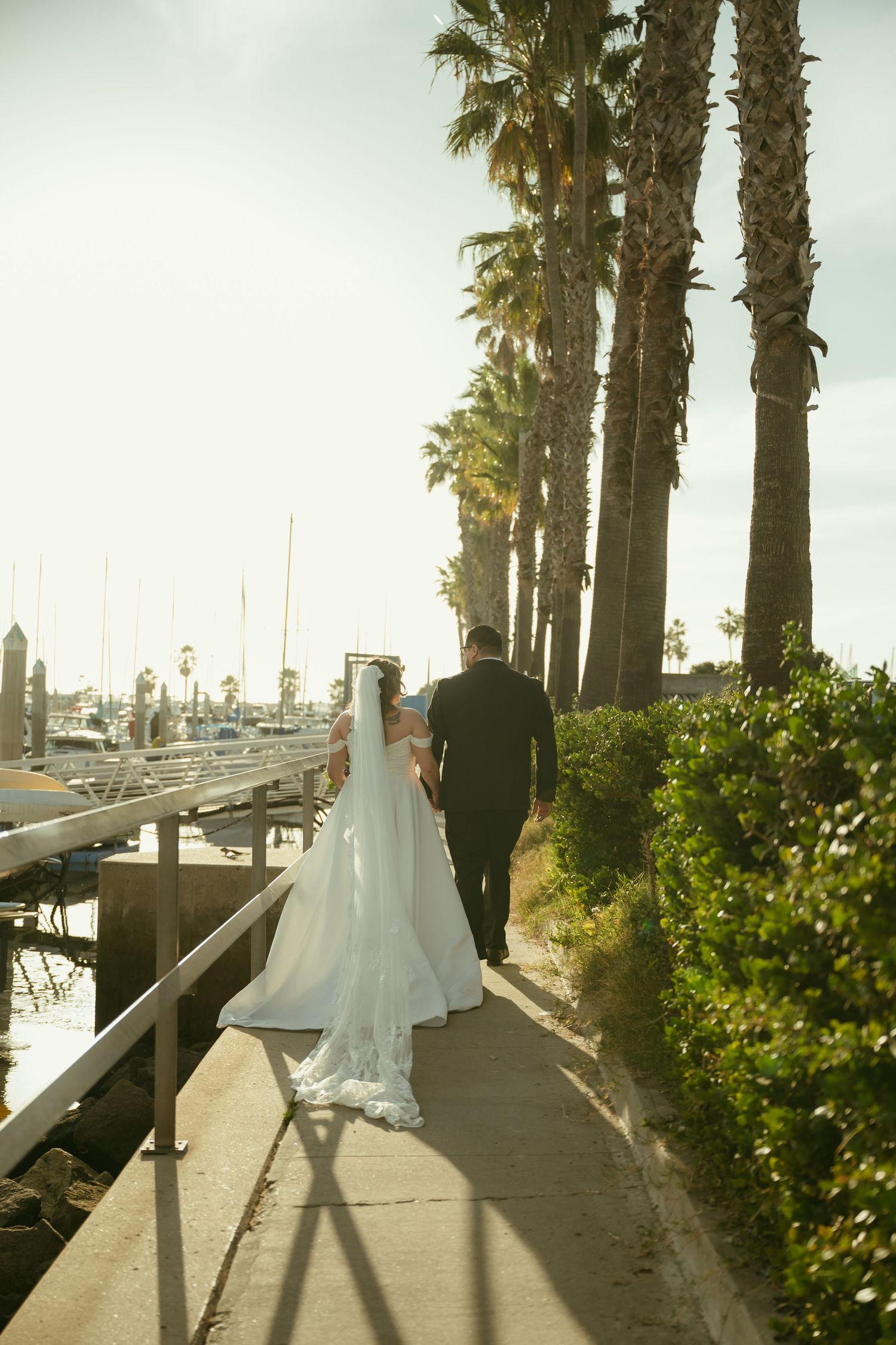 Bride and groom walk hand-in-hand along a palm-lined marina path, bride's white gown trailing behind them.
