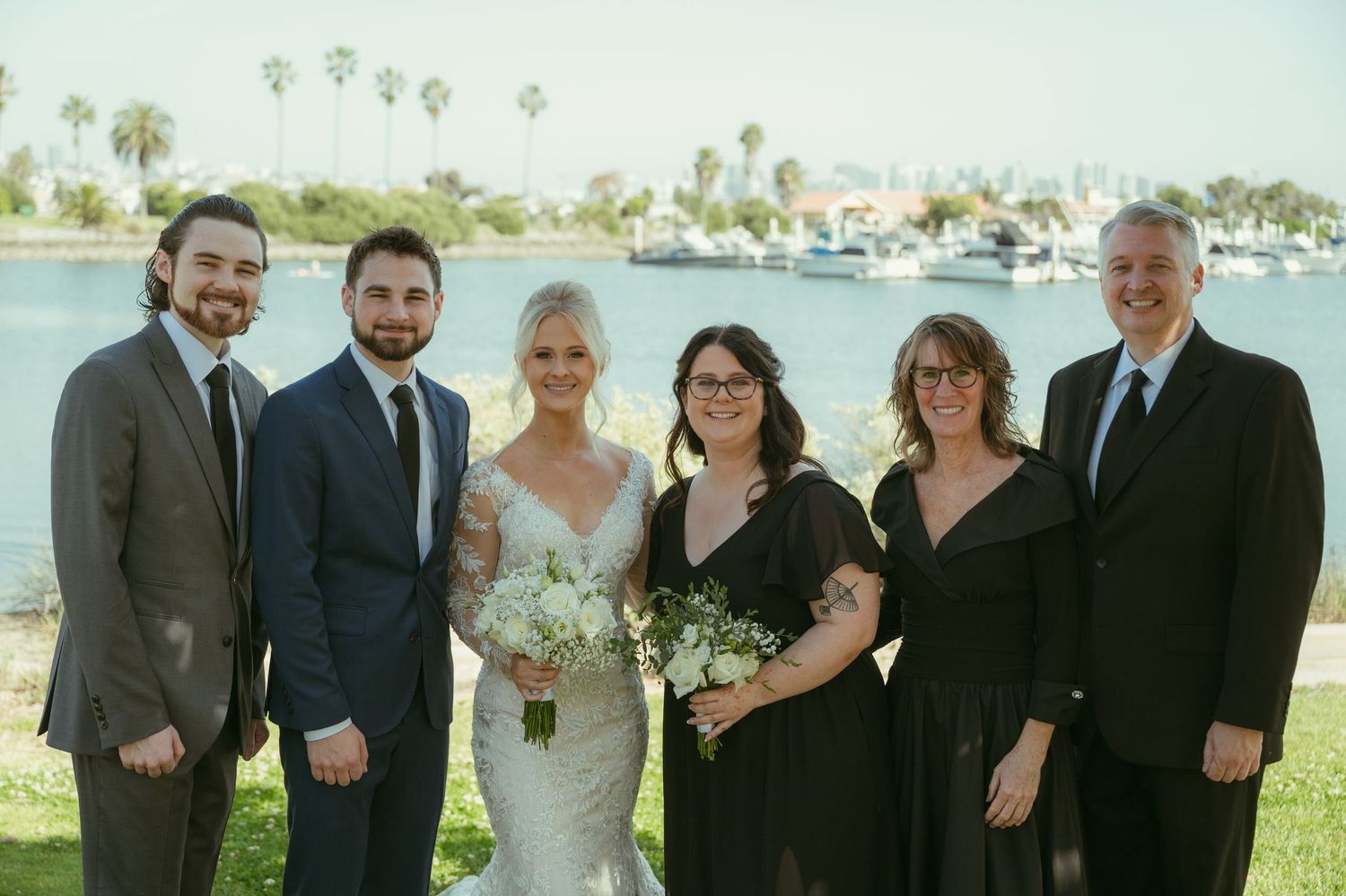 Wedding party poses outdoors by waterfront, bride in lace gown holding white bouquet with family in formal attire.