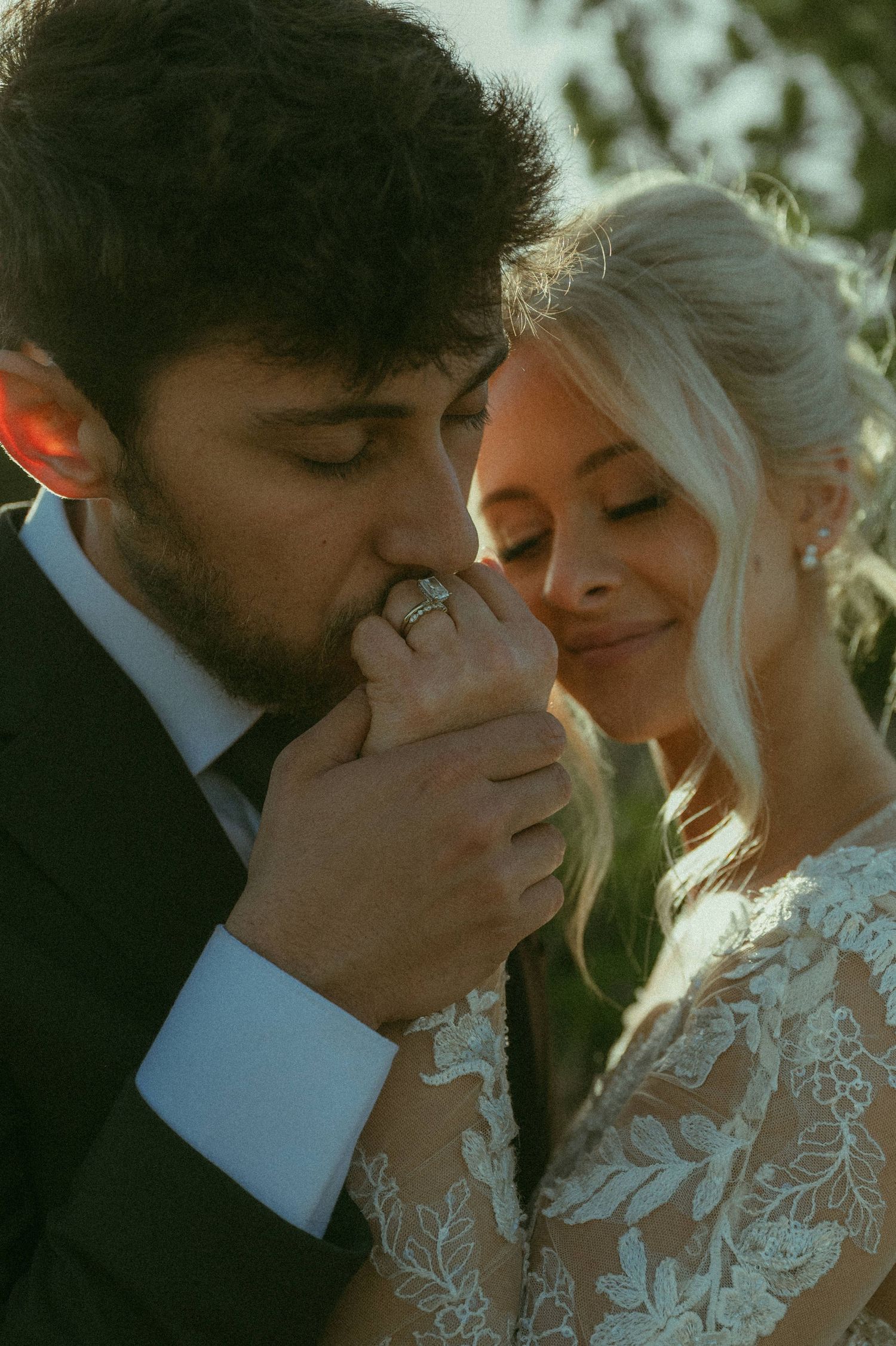 Groom kisses bride's hand adorned with diamond ring while she smiles softly in golden sunset light.