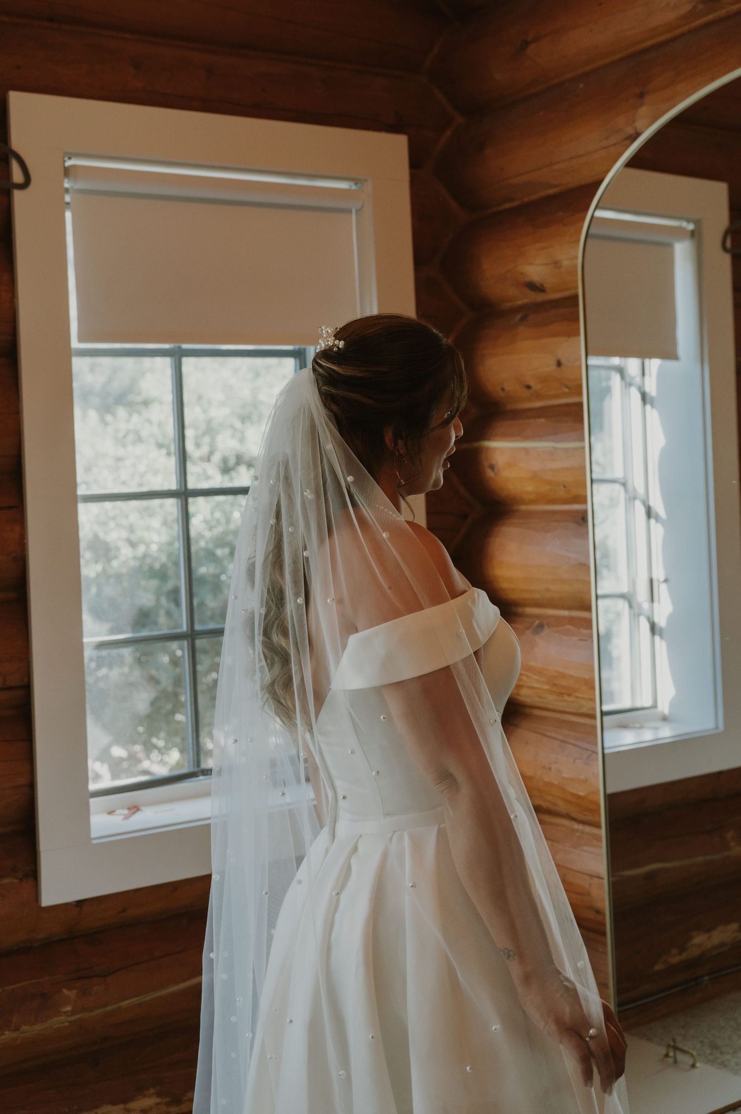 Bride in off-shoulder white gown and pearl veil stands by window in rustic log cabin, viewed from behind.