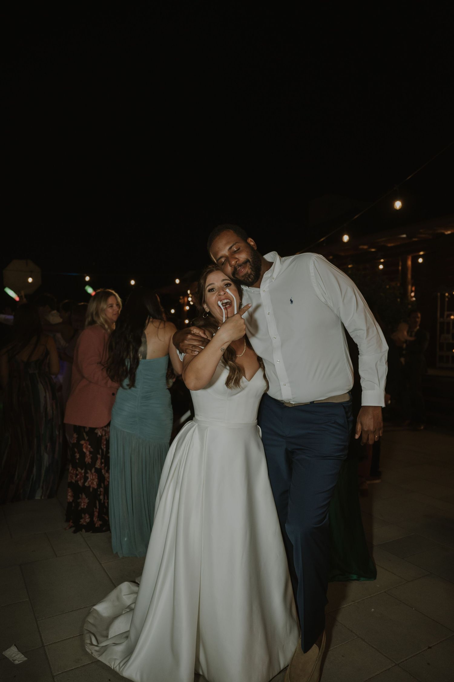 Bride in white gown and groom in white shirt dance joyfully at outdoor wedding reception under string lights.
