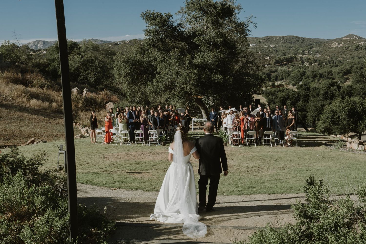 A bride and groom walk hand-in-hand toward their outdoor wedding ceremony with guests seated on a scenic hillside.