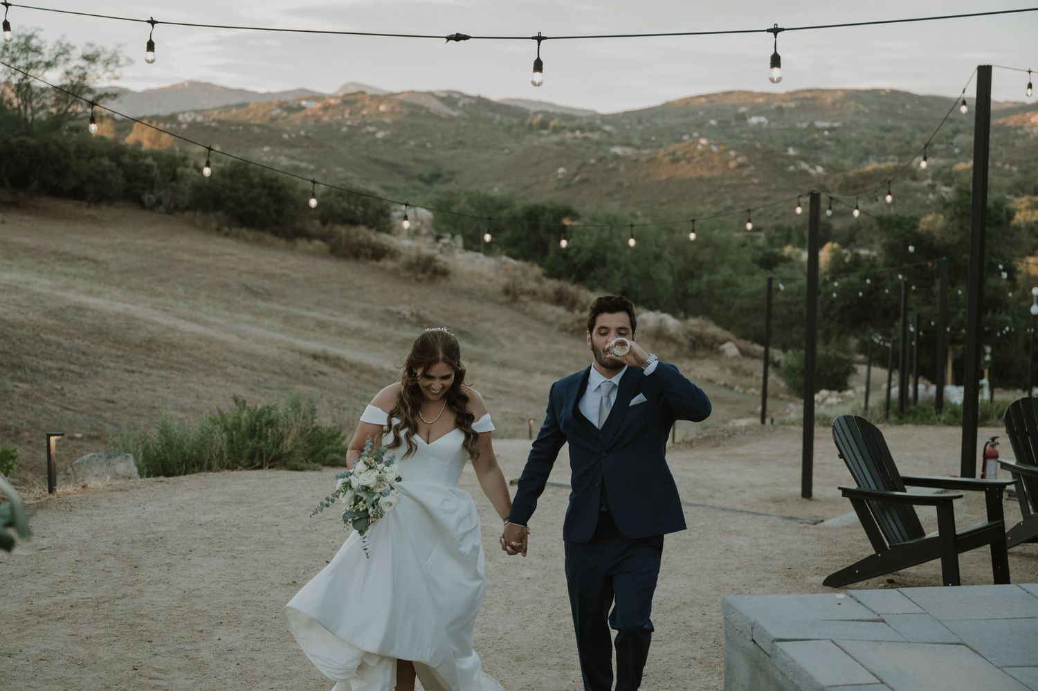 Bride and groom holding hands walking outdoors at a hillside wedding venue with string lights at dusk.