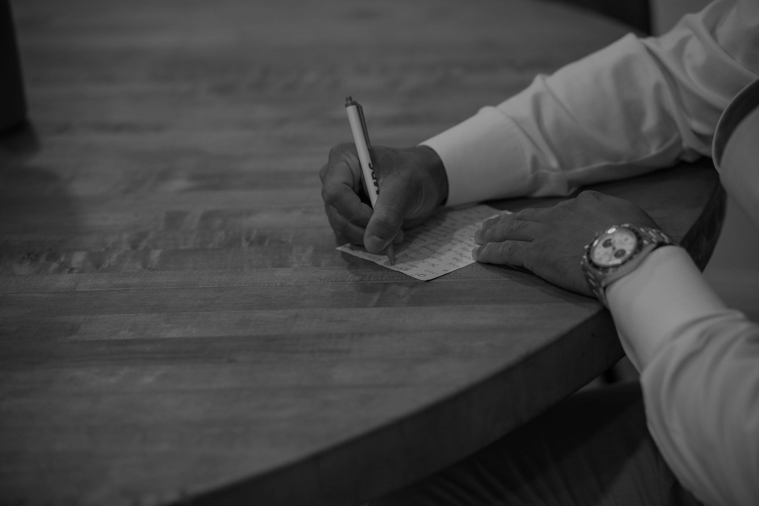 Close-up black and white photo of groom writing wedding vows at a wooden table wearing a watch.